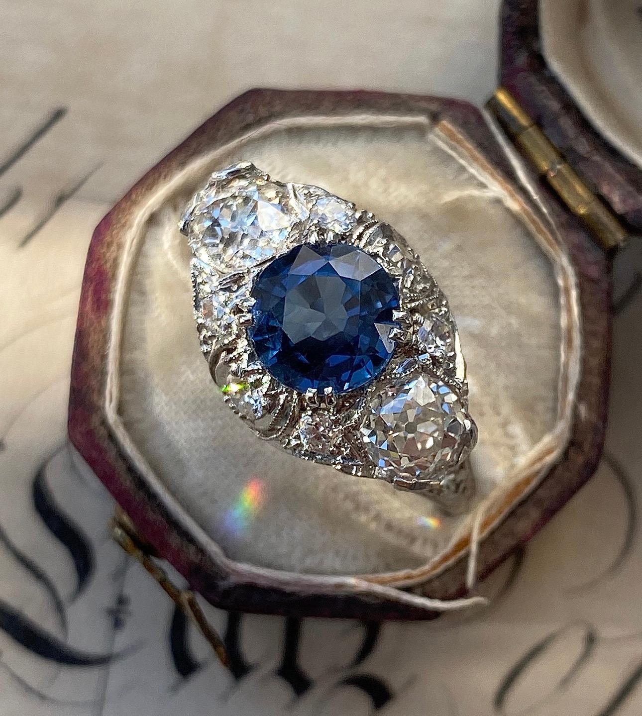 This early 20th century Edwardian/Art Deco ring shimmers from all angles. Hand fabricated in platinum, this lacy diamond setting boasts a 1.64 carat natural, cornflower blue sapphire seated between two sparkling old cut diamonds, skillfully hand
