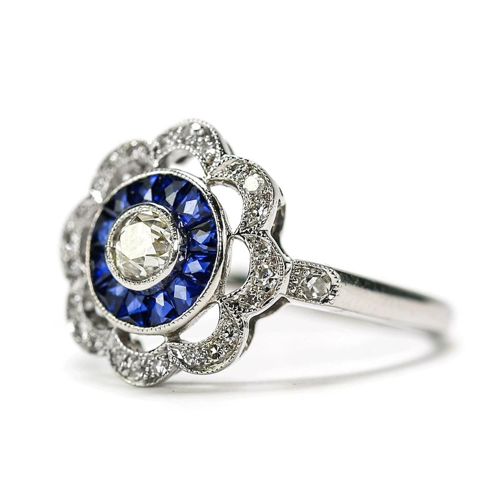 An unusual period 18k white gold ring dating from the Art Deco era with a transitional cut centre diamond est. 0.25cts surrounded by 12 calibre cut sapphires which in turn are faceted too. The scalloped outer rim is mille-grain set with 26 old cut