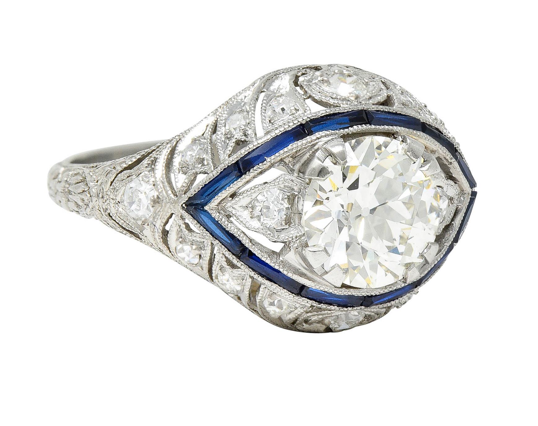Featuring an old European cut center weighing approximately 1.35 carat - J/K color with SI1 clarity

Surrounded by a halo of calibrè cut natural and synthetic sapphires - bright royal blue in color

Bombè style mounting with ribbon-like tendrils and