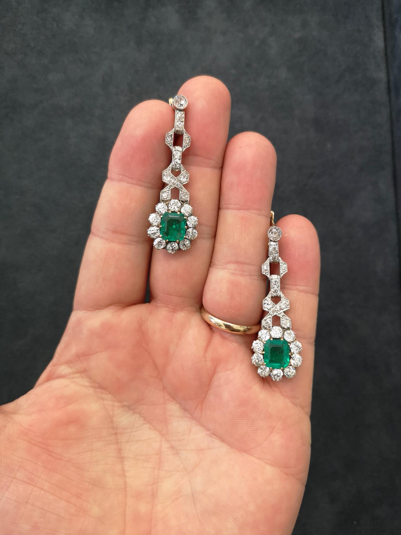 From the much sought after Art Deco period, a pair of 18-carat gold and platinum pendent earrings. Each mounted with a luscious green emerald-cut Colombian emerald of approximately 2.00 carats, surrounded by 10 old-cut diamonds to form a cluster.