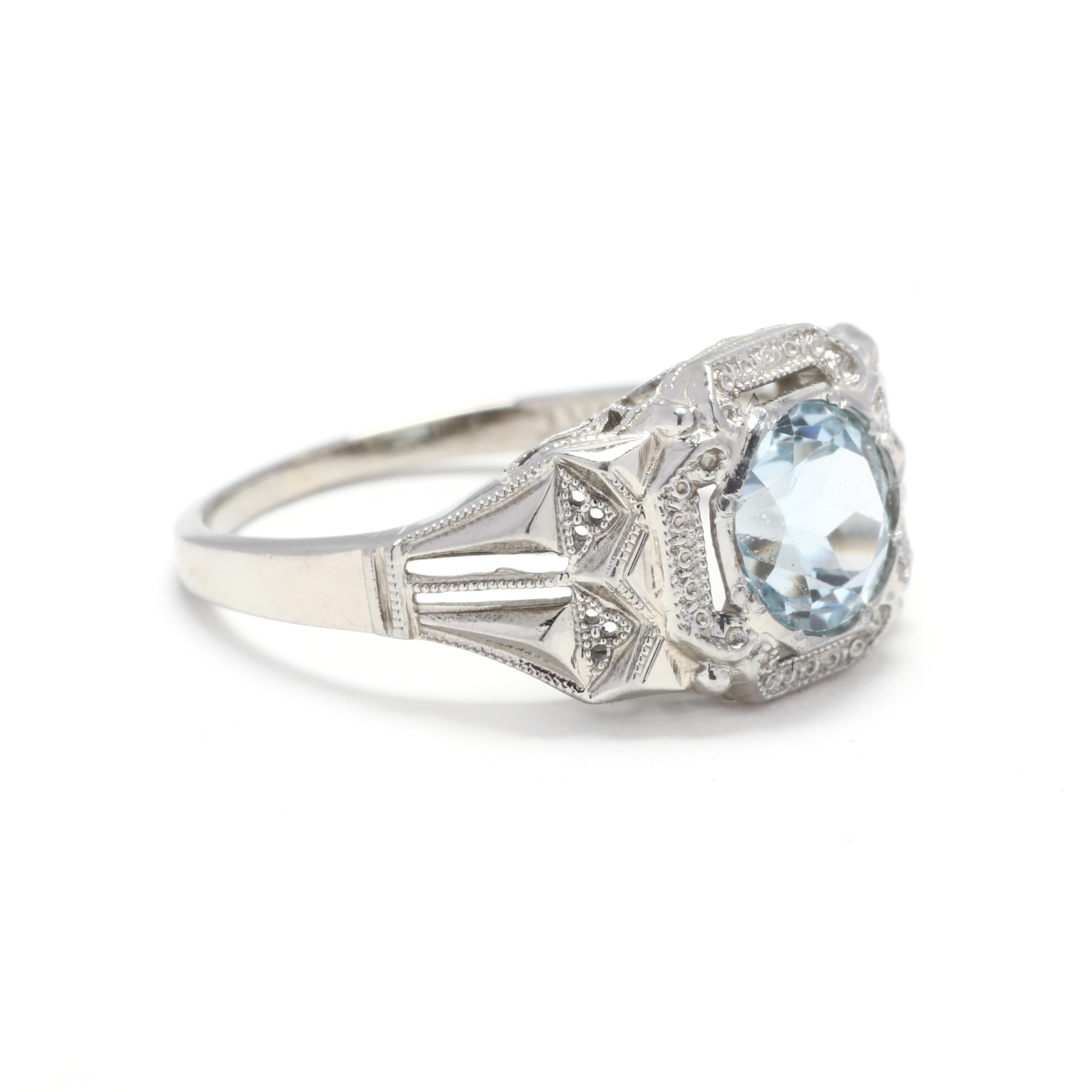 An Art Deco 18 karat white gold and blue topaz engagement ring. This ring features a round cut blue topaz weighing approximately 1.05 carat set in a square scrolled mounting with floral engraving and milgrain detailing.

Stones:
- blue topaz, 1