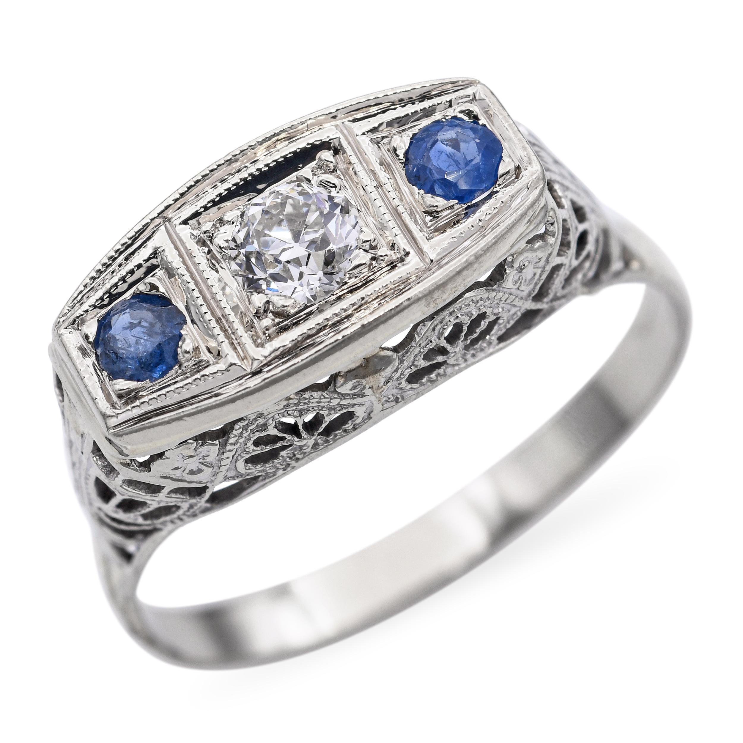 Cette exquise bague art déco est fabriquée en or blanc 18 carats et présente un trio classique avec deux saphirs bleus vibrants flanquant un diamant étincelant de taille européenne au centre. Le diamant présente le charme et le caractère de