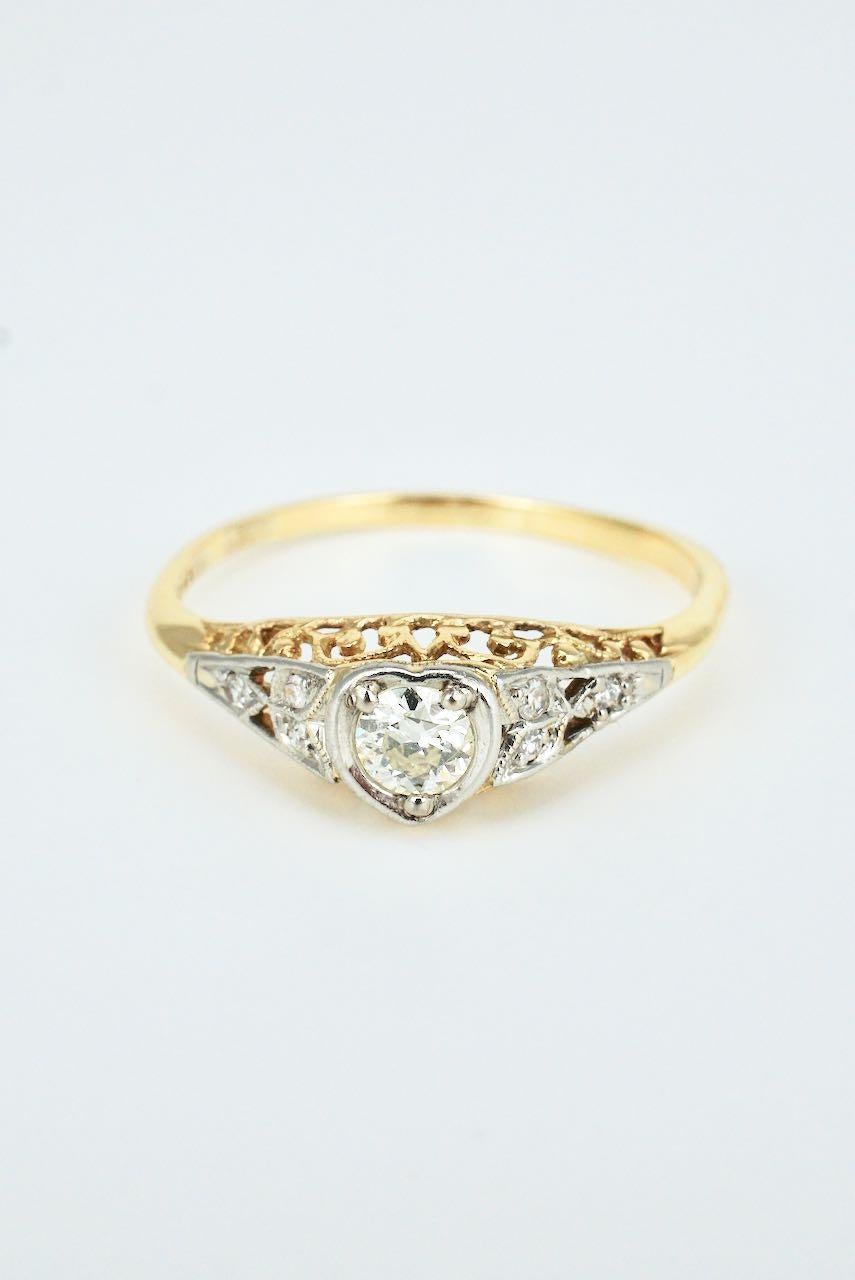An Art Deco heart motif ring featuring a round old European cut diamond set in a heart shape mount with three small round early single cut diamonds to each shoulder set in leaf motifs in platinum above a fretted and millgrained gallery and 18k
