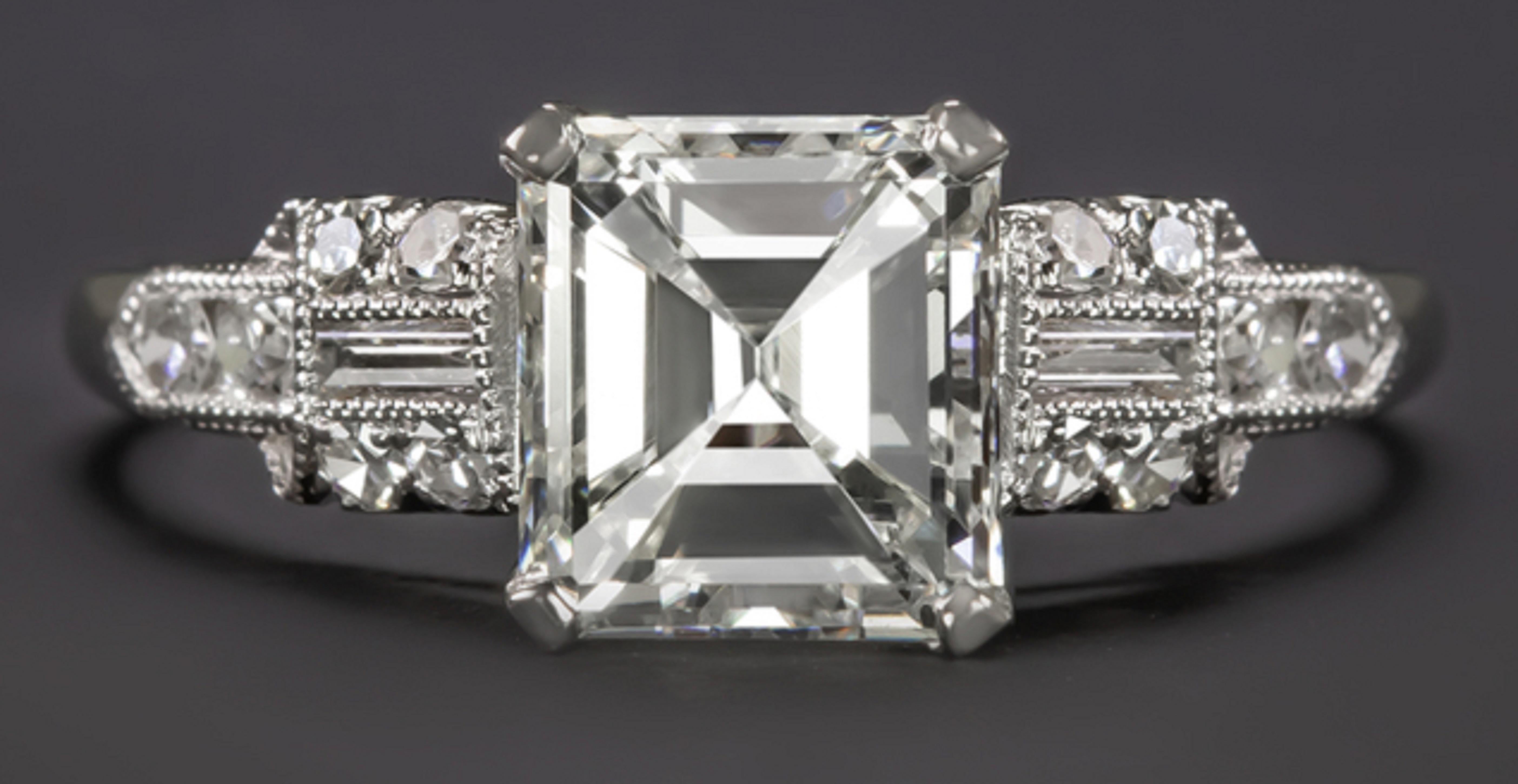This stunning vintage style engagement ring features a vibrant GIA certified emerald cut diamond complemented by an elegantly designed diamond studded setting. The main stone is a very white white and completely eye clean 1.30ct center diamond an