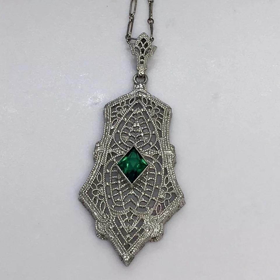 Questa splendida collana è un meraviglioso esempio di stile Art Déco. Il pendente presenta un bellissimo cristallo verde incastonato in un castone, circondato da diamanti più piccoli. La collana è realizzata in oro bianco 10k e presenta un design in