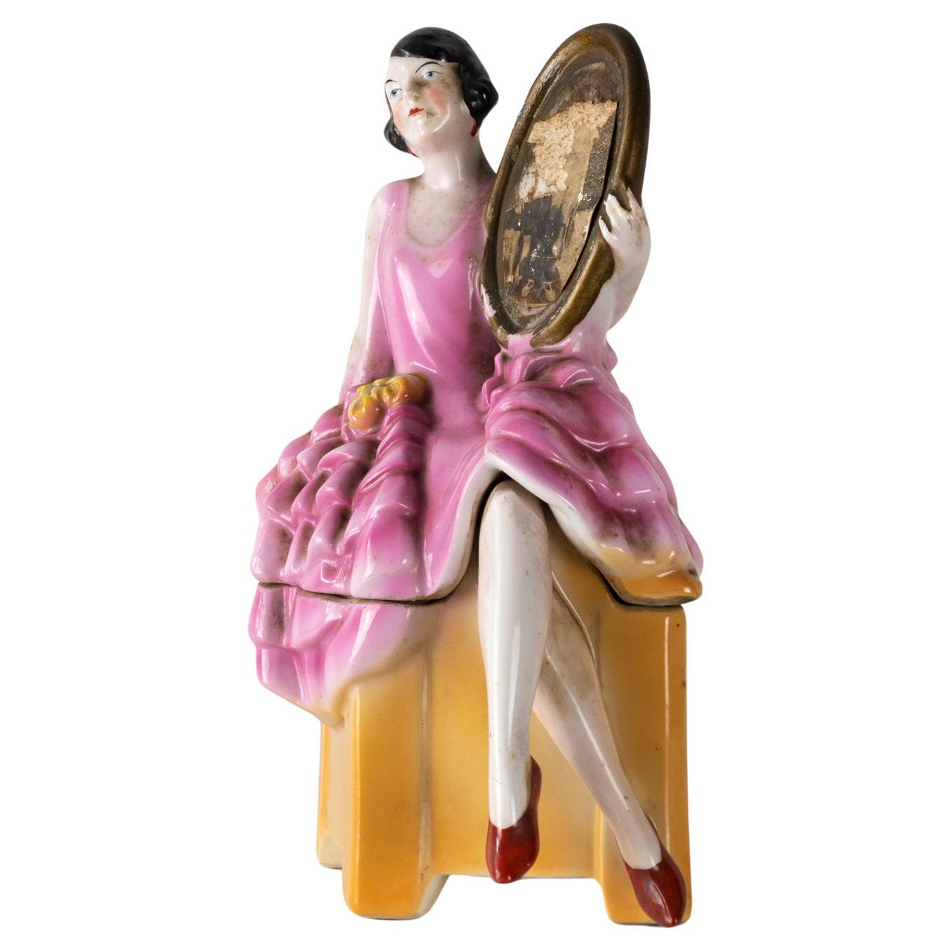 Deutsche Keramik-Pulverdose aus dem Art déco der 1930er Jahre /  Schmuckkästchen in Form einer Art-Déco-Hutschachtel mit einem Flapper-Girl, das auf dem Deckel sitzt und einen Spiegel hält.   
Wunderschöne Färbung und Detailtreue.
Ein seltenes