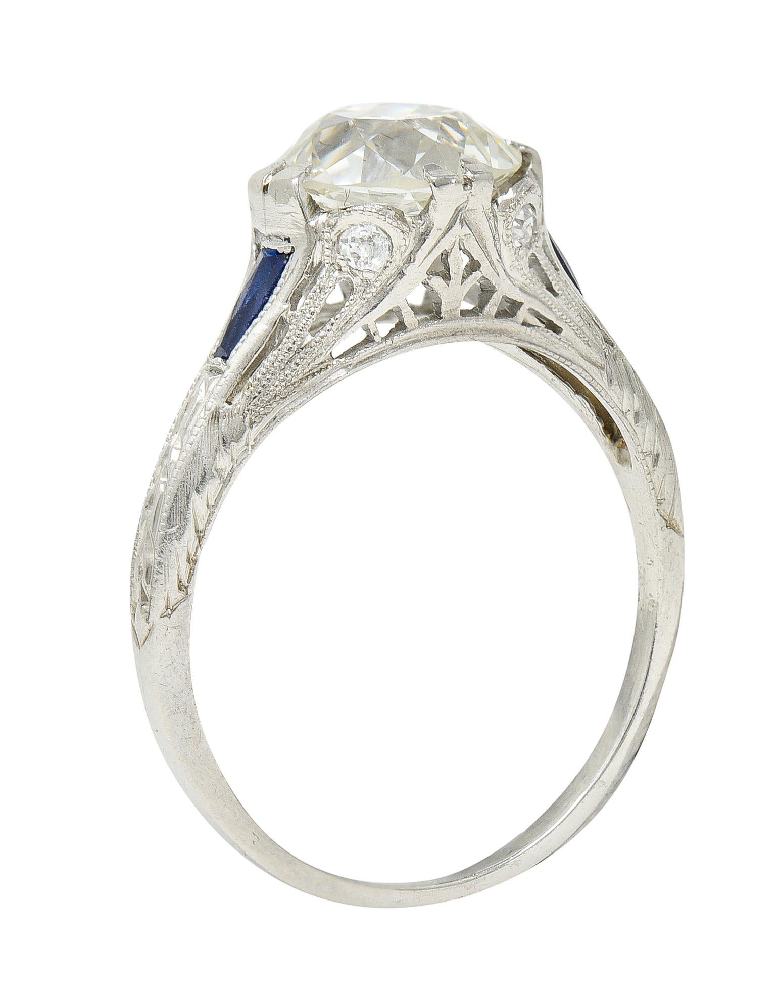 Featuring an old European cut diamond weighing 1.85 carat with K color and VVS2 clarity. Set by wide split prongs in a decoratively pierced mounting with milgrain details. Flanked by tapered baguette synthetic sapphires - well matched and bright