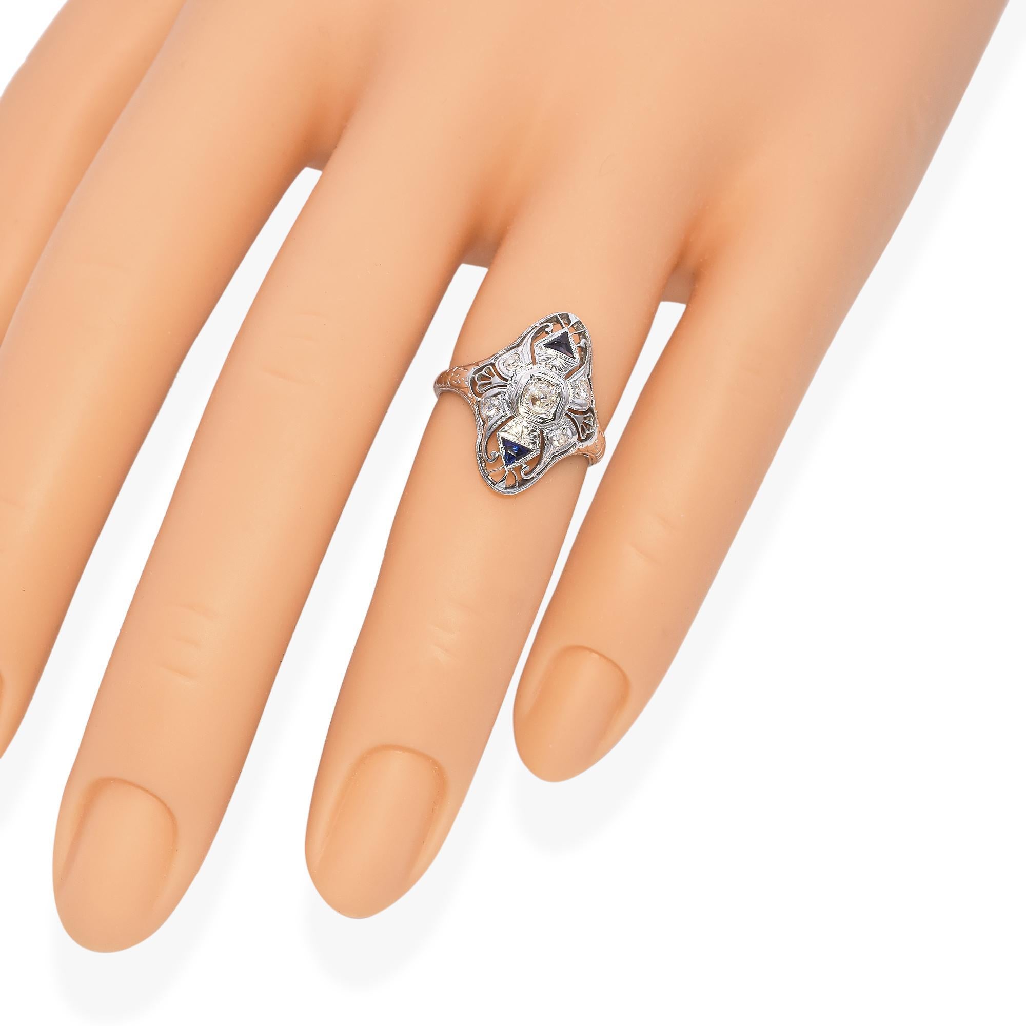 This intricate ring is finely crafted in 19k white gold. The ring features a central .11ct old mine cut diamond, surrounded by delicate filigree work. Two triangular blue sapphires accentuate the design, adding a touch of color. The frame is