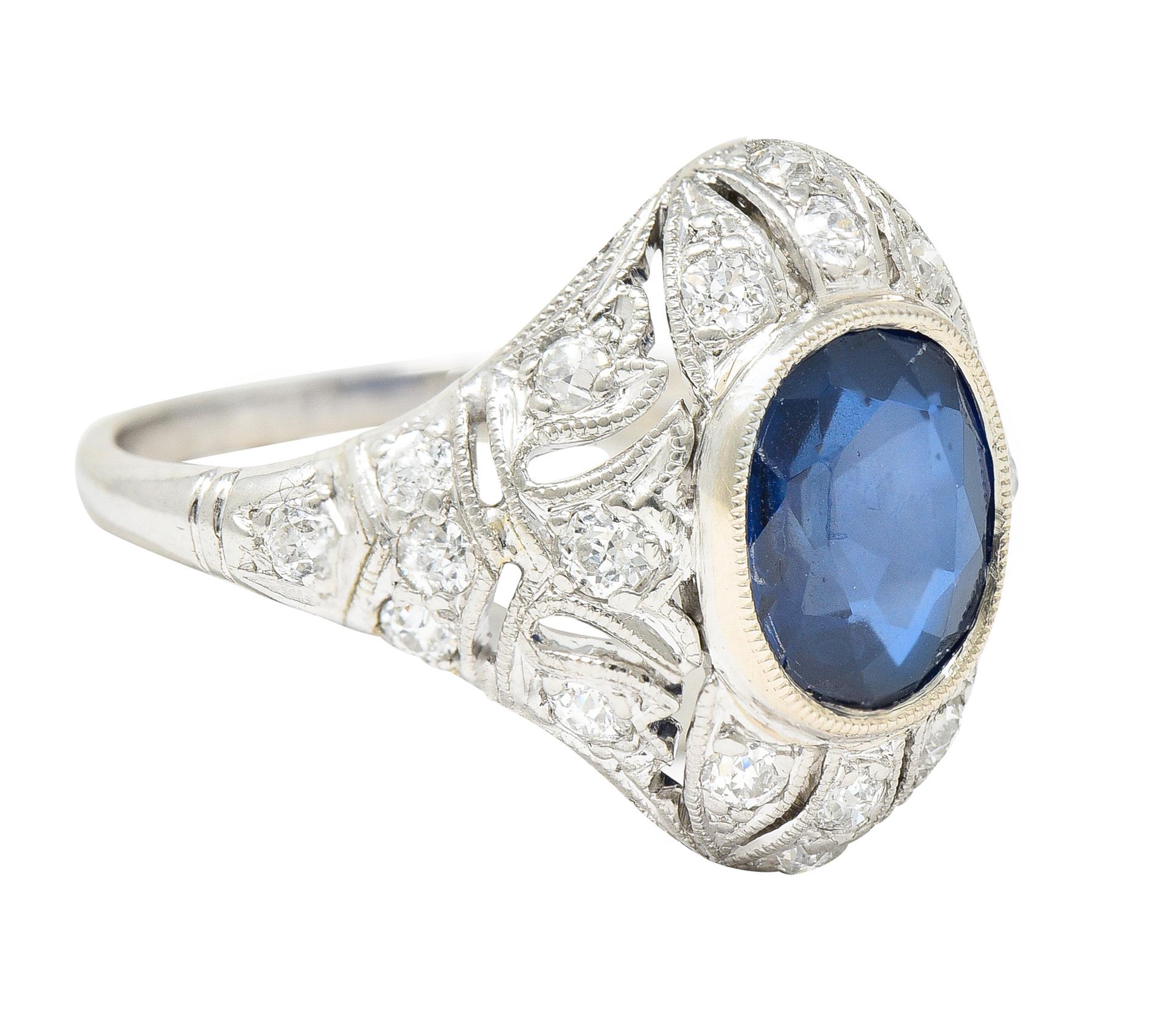 Centering a cushion cut sapphire weighing approximately 1.62 carats total - transparent medium/dark royal blue. Set in a milgrain bezel with a pierced scrolling foliate motif surround with single cut diamonds. Bead set and weighing approximately