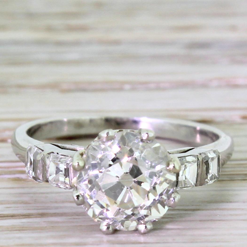 A mega and magnificent Art Deco treasure. This big, bright and vibrant old mine cut stone rests in a triumphant platinum setting. The central solitaire holds a soft champagne hue, deliberately emphasised by the four colourless asscher cut diamonds