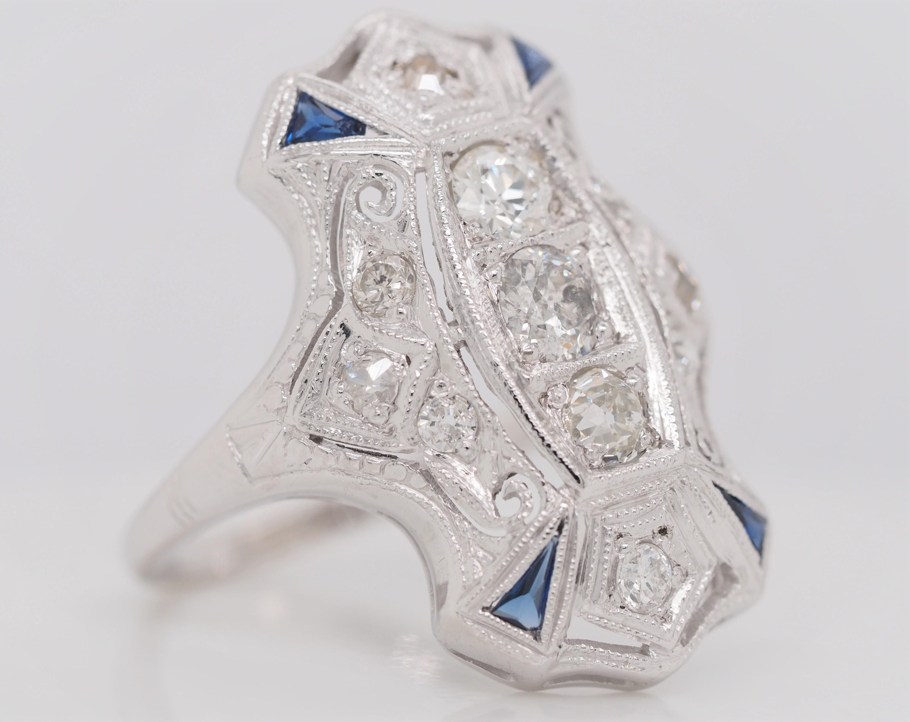 This lovely example of an Art Deco shield ring is crafted of stunning platinum. Intricate filigree designs surround 3 sparkling diamonds on this vintage beauty. Four sapphire pyramids and 8 round cut diamonds add a pop of color that adds to this