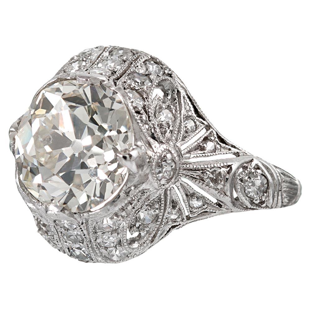 Hand made in platinum in the grand art deco tradition, the ring contains a 3.03 carat center round brilliant diamond that exhibits K color with Si1 clarity. The stone is enshrined in a finely detailed and ornate mounting with a bow motif on each