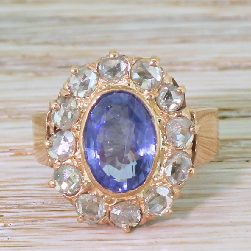 An impressive vintage sapphire and diamond ring. The oval cut sapphire in the centre displays a mesmeric, bright blue, and is secured in a rose gold migrained rubover setting. The centre stone is surrounded by twelve rose cut diamonds in a coronet