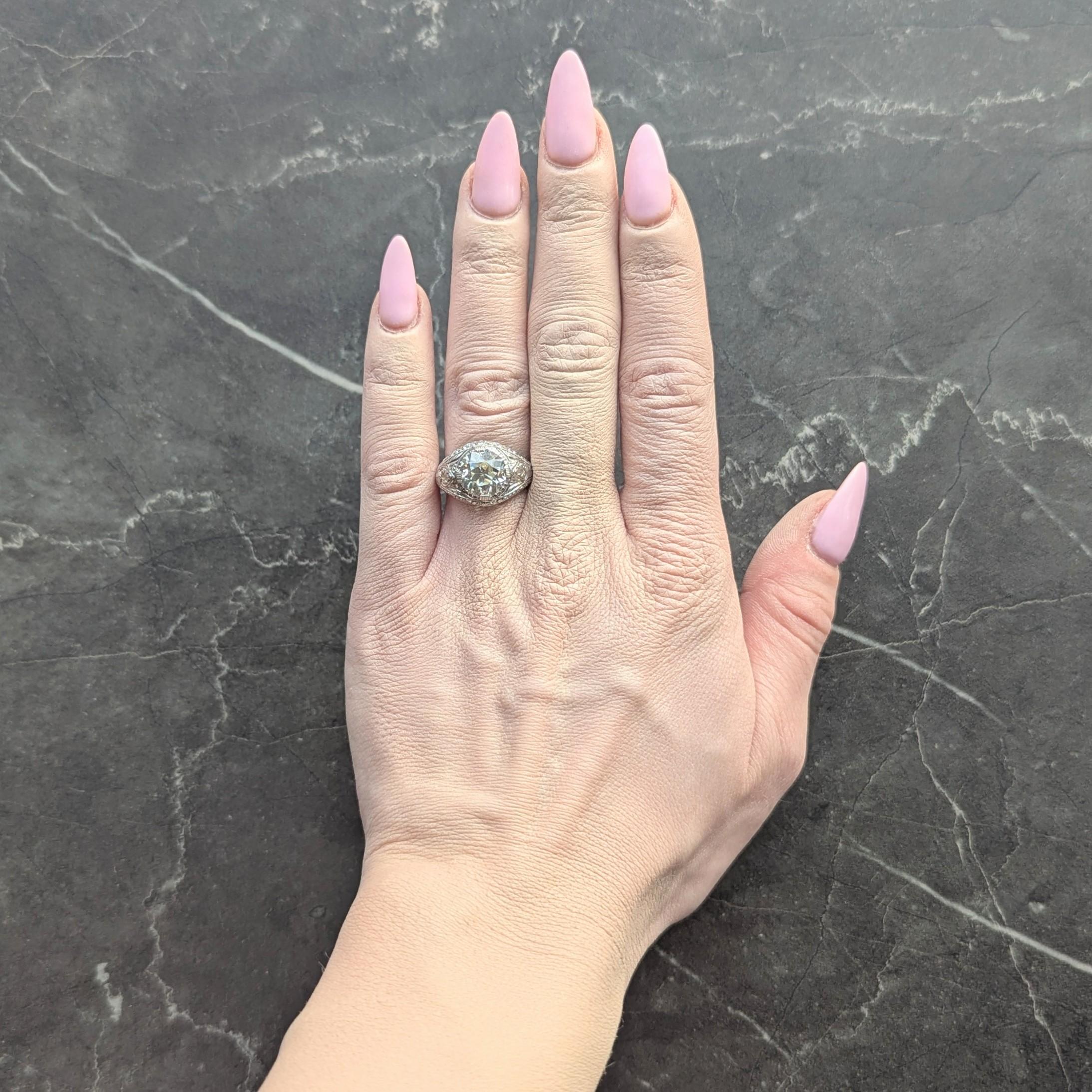 Centering an old European cut diamond weighing approximately 3.07 carats
M color with VS2 clarity - set wit h grooved wide prongs in bombé surround
Engraved with fanning palm leaf and lotus motif shoulders
Accented by scroll motif borders continuing