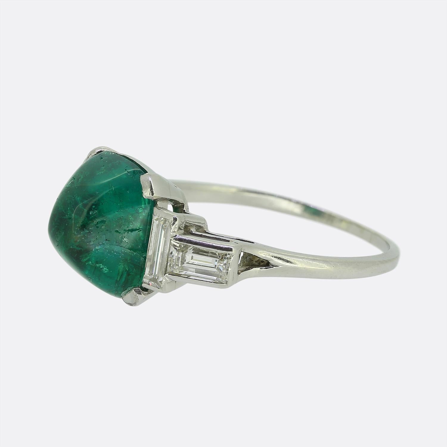 Aquí tenemos un llamativo anillo Art Déco elaborado en platino. En su corazón yace una cautivadora esmeralda talla cabujón, que emana un rico y vibrante tono verde. Flanqueando esta piedra focal a ambos lados hay dos diamantes rectos de talla