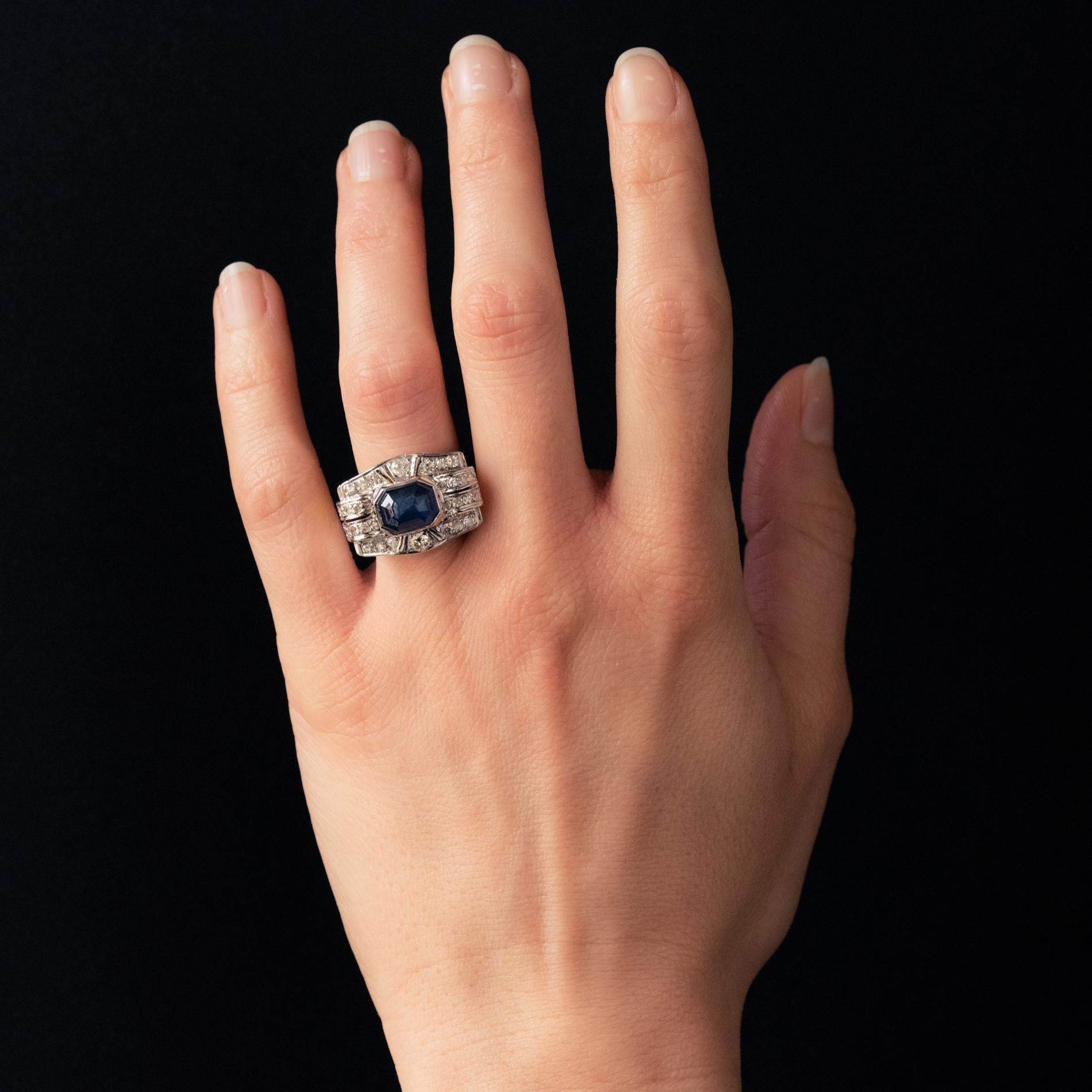 Ring in platinum, mascaron hallmark.
Rectangular and wide, this sublime art deco ring is adorned on top with a faceted rectangular- cut cabochon blue sapphire. The whole tour is paved with diamonds. The basket is perforated with geometric