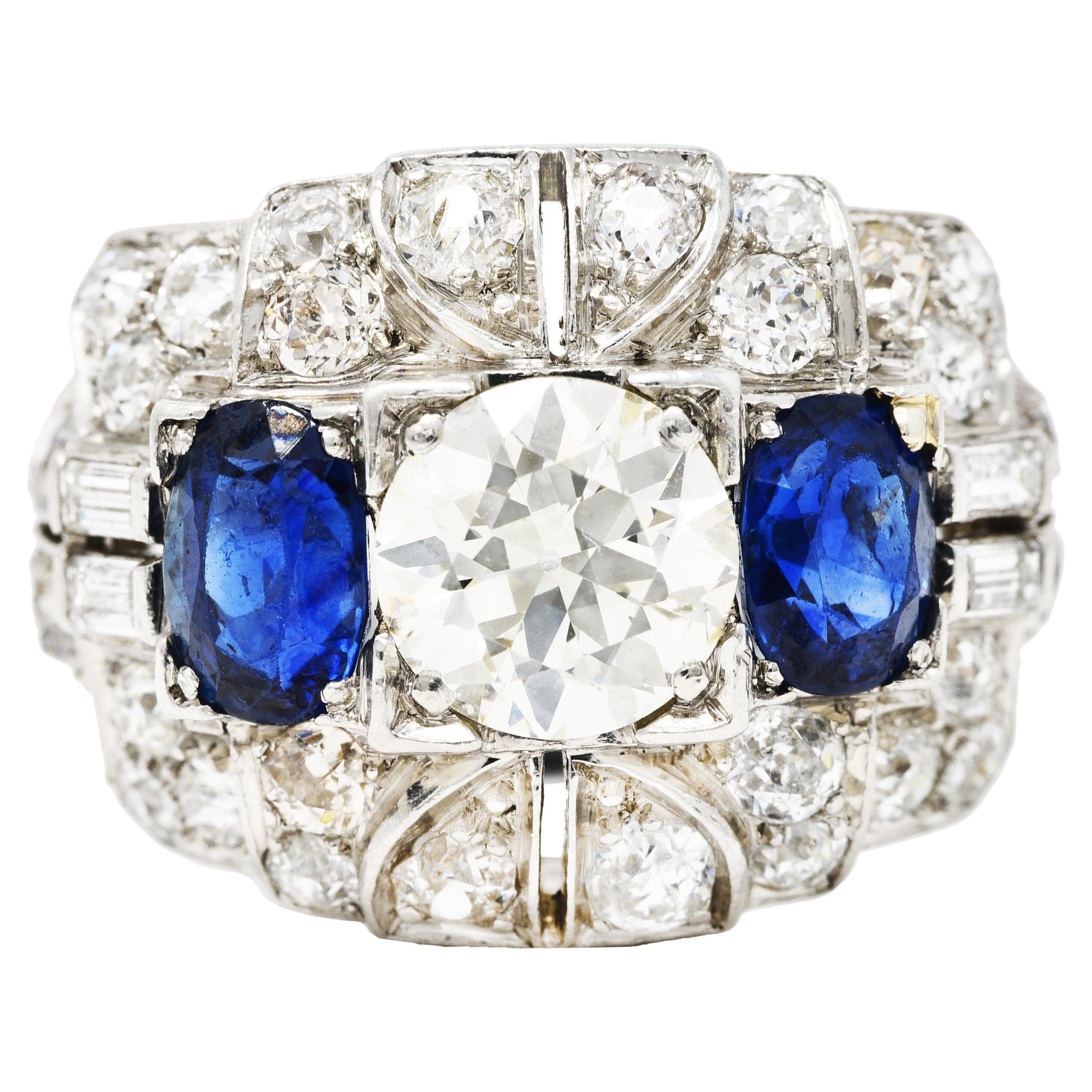 Domed bombè band ring is comprised of undulating geometric forms. Centering an old European cut diamond weighing approximately 1.61 carats - K color with SI1 clarity. Set low in a square form head and flanked by two oval cut sapphires. Very well