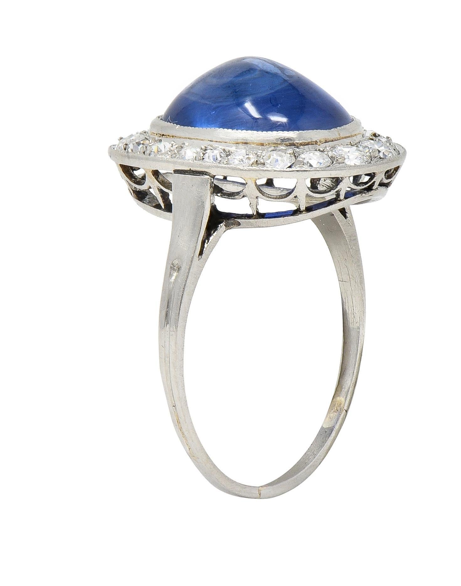 Centering a sugarloaf-shaped sapphire cabochon weighing 6.12 carats - transparent medium blue 
Natural Burmese in origin with no indications of heat treatment - bezel set
With a halo surround of old European and transitional cut diamonds 
Weighing