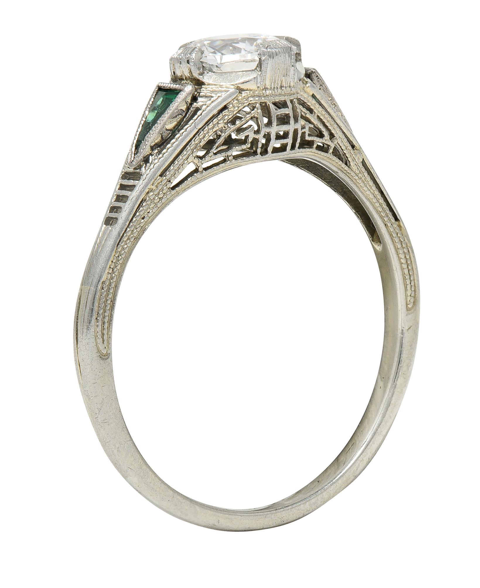 Centering an old European cut diamond weighing .65 carat - G color with VS2 clarity 
Set with compass-style tab prongs flanked by two triangle cut synthetic emeralds
Weighing approximately .06 carat - bezel set 
Featuring a pierced Greek key motif