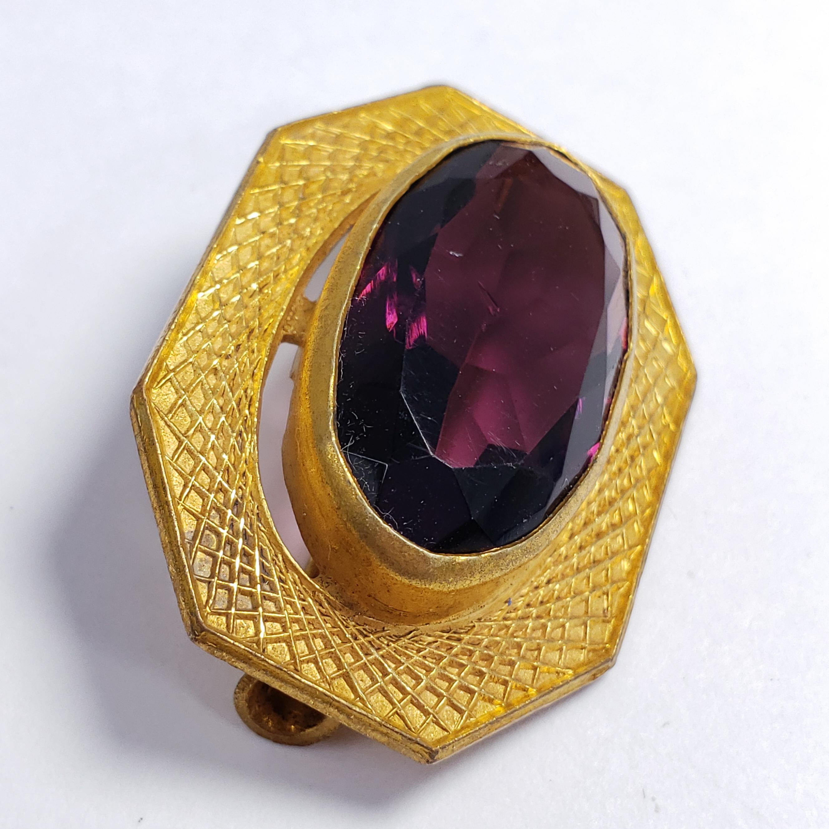 An elegant art-deco pin brooch, featuring an amethyst-colored open-back crystal centerpiece in a goldtone bezel. Features a vintage-style pin hook, typical of the era.