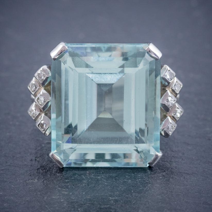 An extravagant Art Deco cocktail ring adorned with a magnificent emerald cut aquamarine with a calming clear blue hue with flashes of green, weighing an impressive 25 carats (approx.) 

Aquamarine derives its name from its calming, clear blue/ green