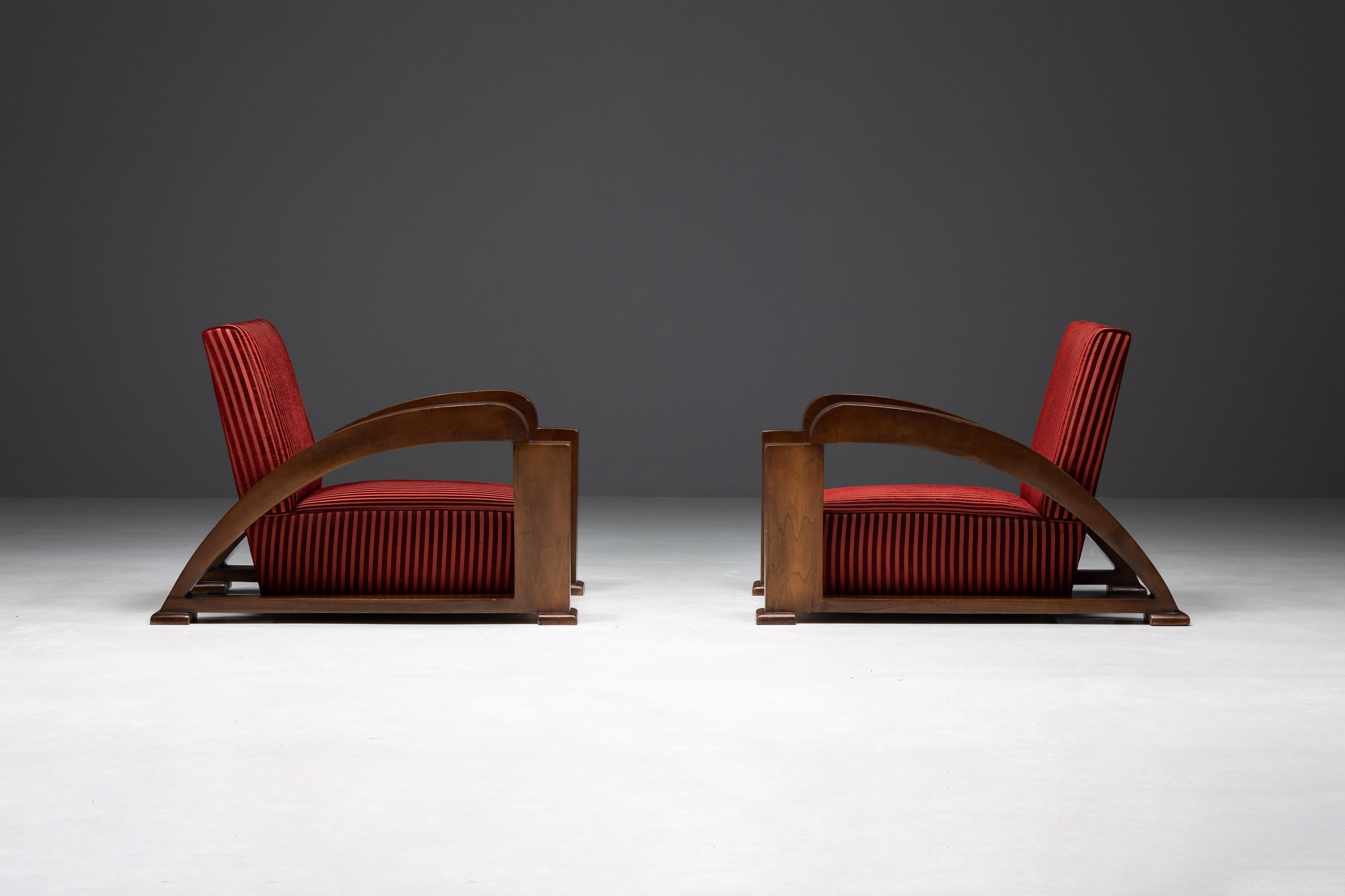 A pair of stunning Art Deco armchairs that encapsulate the sophistication of the early 20th century. These lounge chairs boast gracefully curved armrests, adding an air of elegance to their substantial presence. The fruitwood veneered frames exude