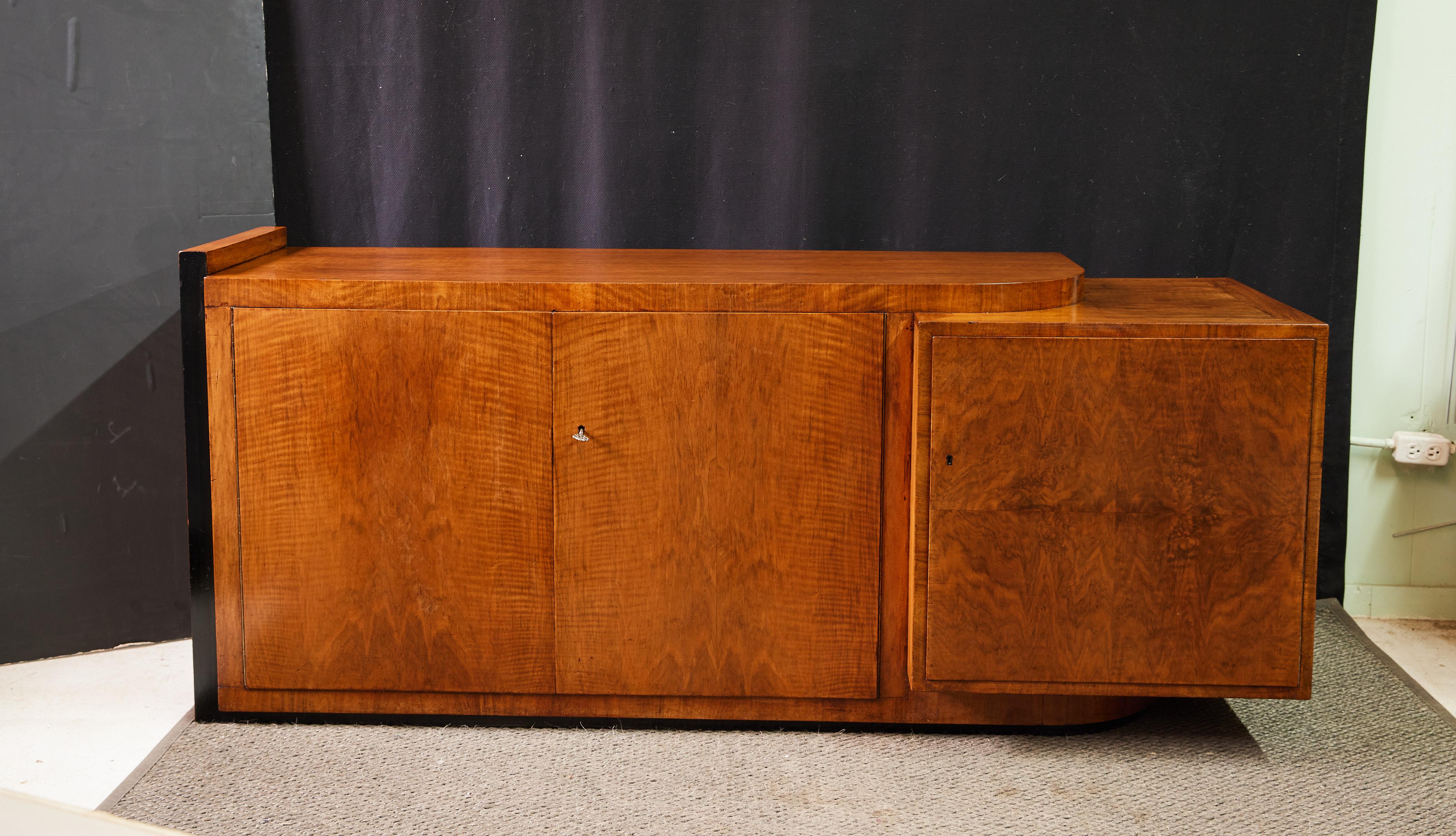 Early 20th century Art Deco period credenza of an impressive asymmetrical form and veneered in burl walnut. The primary case holds a flush cabinet with double doors that open to a storage cavity with a wide shelf below a pair of felt-lined drawers