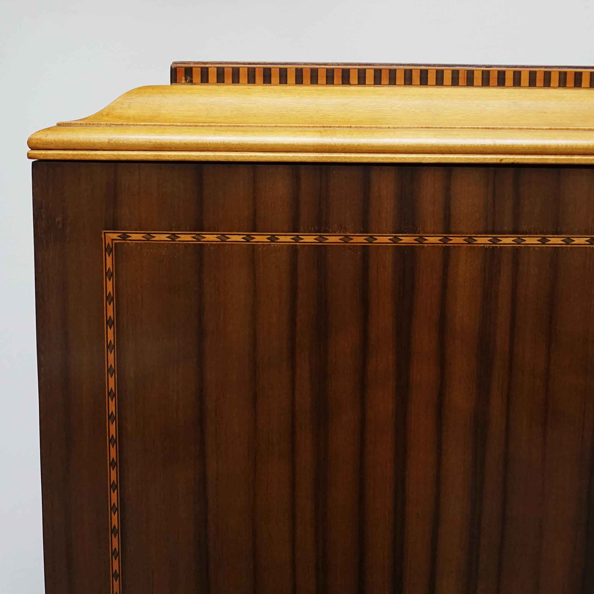 An Art Deco cabinet on tapered legs. Upper cabinet doors with two ivorine vases as decoration open to reveal interior shelving and a lower pull out drawer. Australian walnut veneered with ebonised and satinwood inlay and original brass handles with