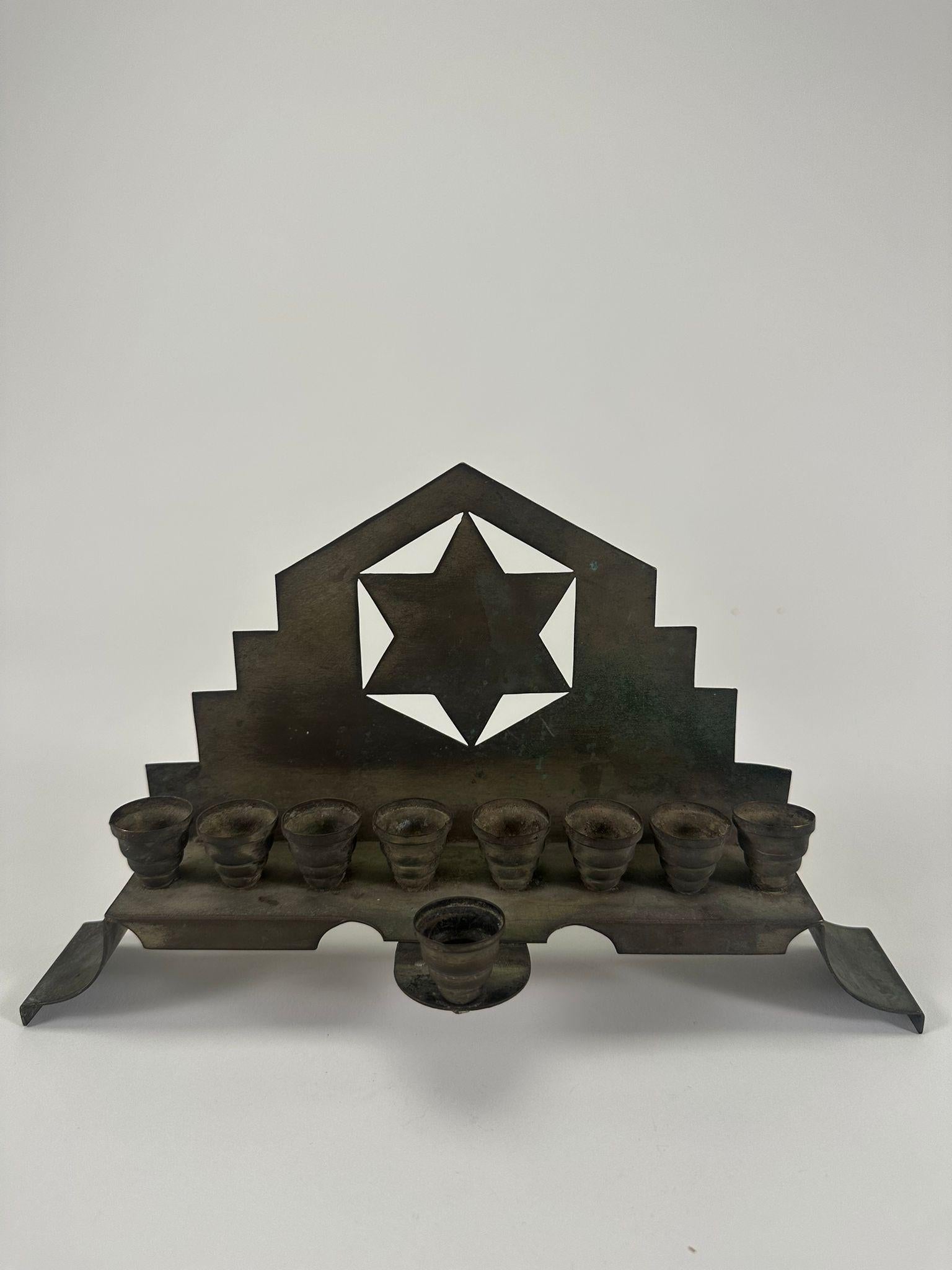 Questa straordinaria menorah in rame Art Deco, realizzata in Austria nel 1925, è una fusione magistrale di estetica modernista e tradizione ebraica. La piastra posteriore della menorah presenta un audace design geometrico a gradini, che ricorda lo