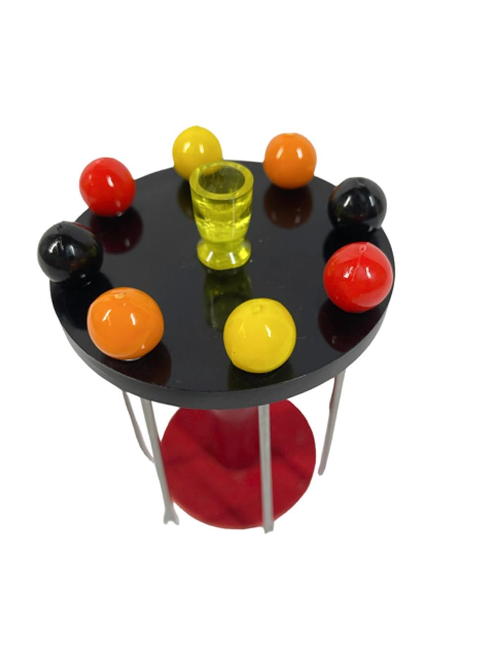 Set of eight cocktail picks and stand, 2 each of 4 color picks in steel with ball finials hang from the top of a table-form stand. The table with a black Bakelite top supported on a red Bakelite column sitting on a red felted disk, A translucent