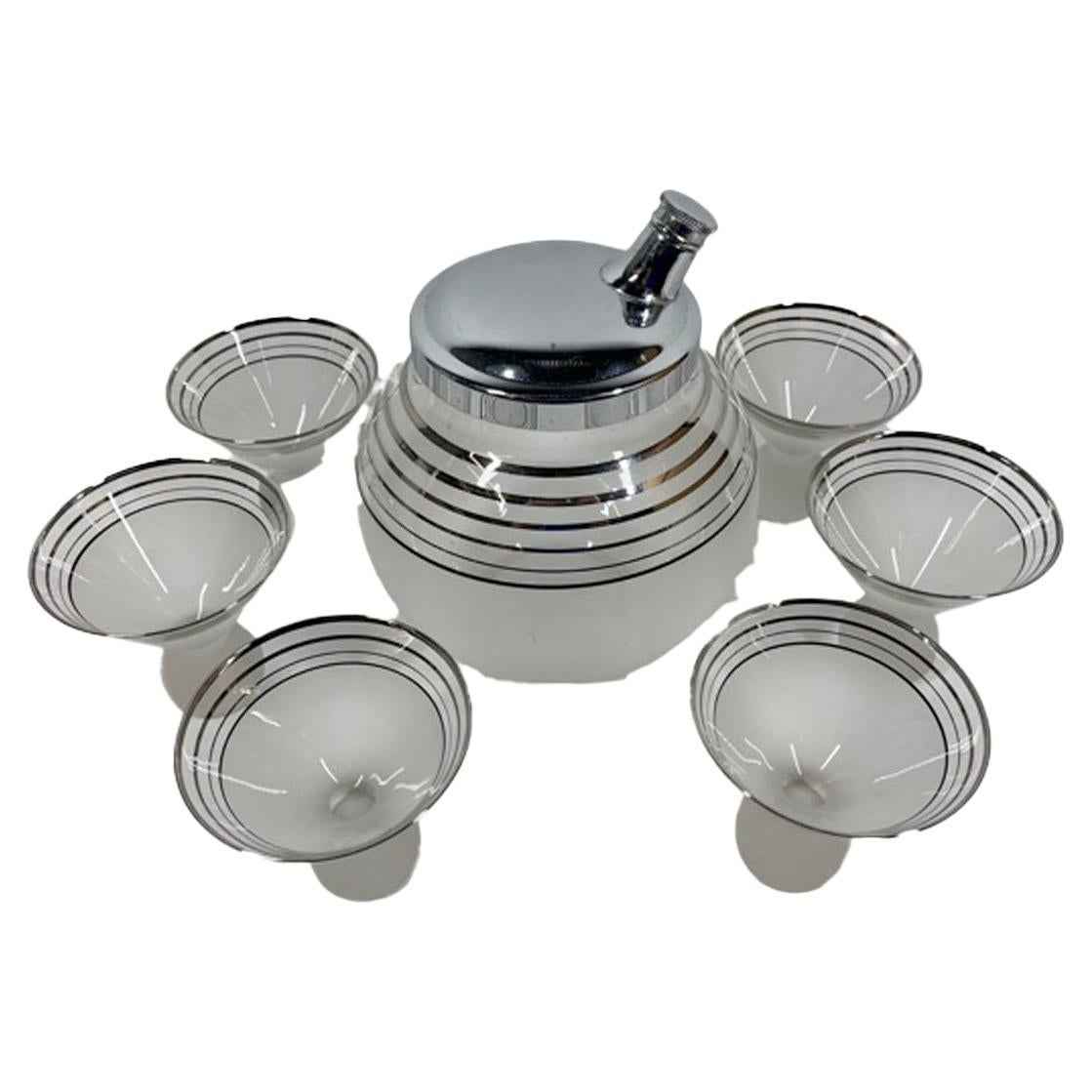 Set da cocktail Art Deco con shaker a forma di palla e 6 bicchieri da cocktail con coppe a forma di cono su piedini a sfera. Ogni pezzo presenta bande argentate su vetro trasparente sopra una sezione inferiore smerigliata.