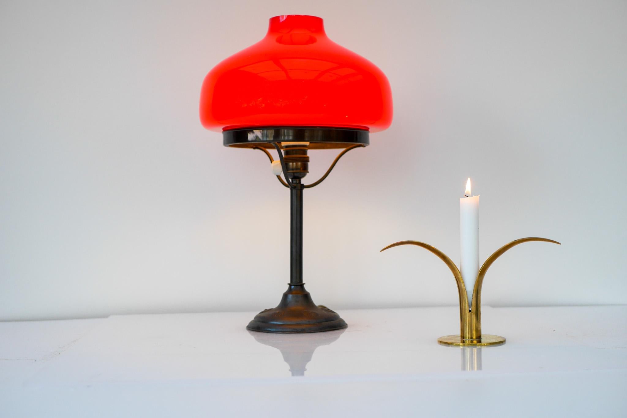 A bright red Banker's metal and glass table lamp from 1930s, Sweden. It has a classic shape with a metal base, with some flower details and arms which holds up the bright red glass dome. The lamp is very elegant and gives a warm orange light once