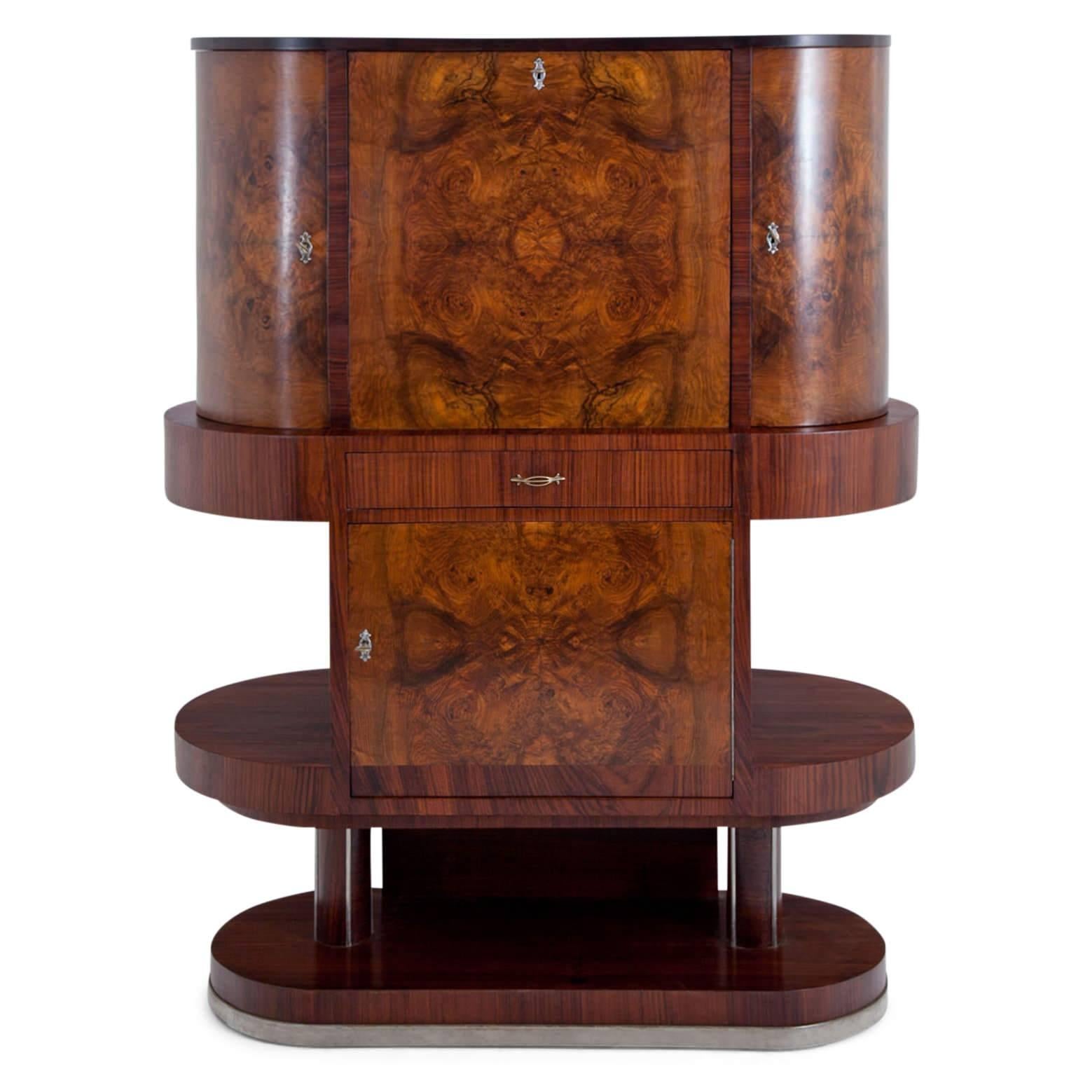 Tall Art Deco Bar Cabinet on an oval stand, with a middle compartment with two shelves and a three-doored top part. The door of the central cabinet folds down and the doors to the sides are rounded. The interior offers space for bottles and glasses.