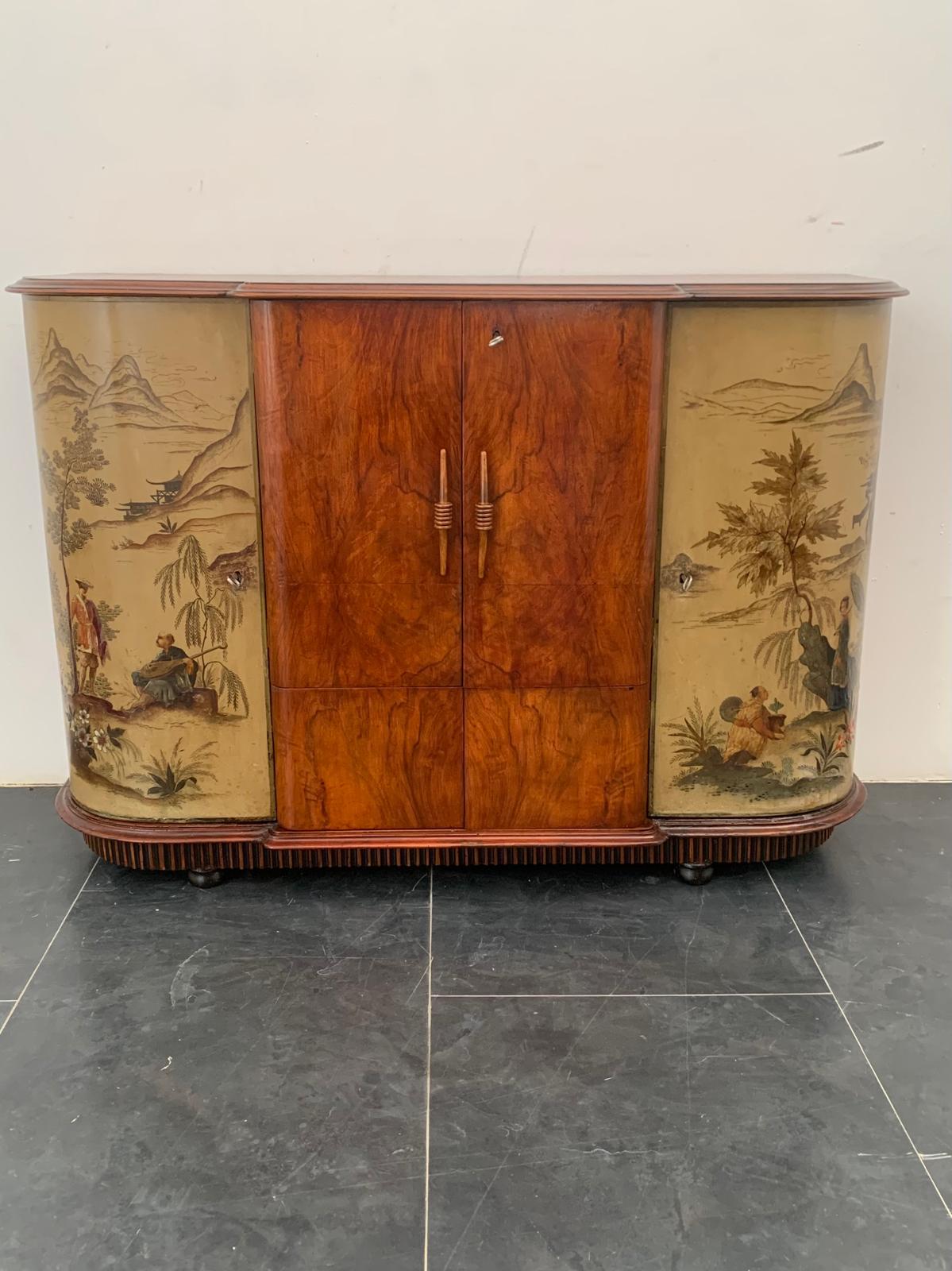 Mobile bar Art déco in noce, la base grissinata o mille righe sostiene il corpo del mobile arrotondato ai lati, al centro una splendida radice di noce con maniglie intagliate racchiude il vano bar. I lati sono finemente laccati e raffigurano scene