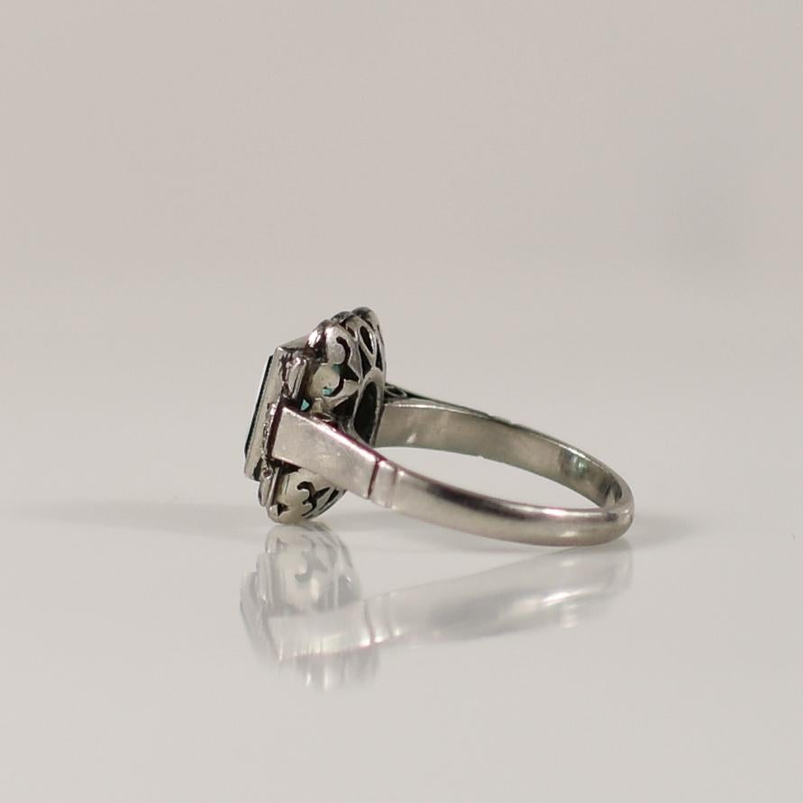Dieser antike Art-Déco-Ring zeigt einen faszinierenden natürlichen Smaragd im Smaragdschliff, der in der Mitte der Lünette gefasst ist. Der Smaragd wird von einer Reihe funkelnder Diamanten umrahmt, die seinen leuchtend grünen Farbton