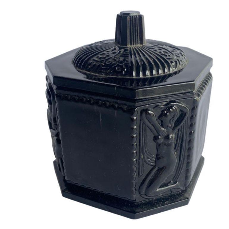 This antique 1920's classical black amethyst hexagon shaped Tiara Glass vanity jar presents a captivating blend of elegance and antiquity. Crafted with meticulous detail, the jar features a Greecian nude woman depicted in classical style, adding a