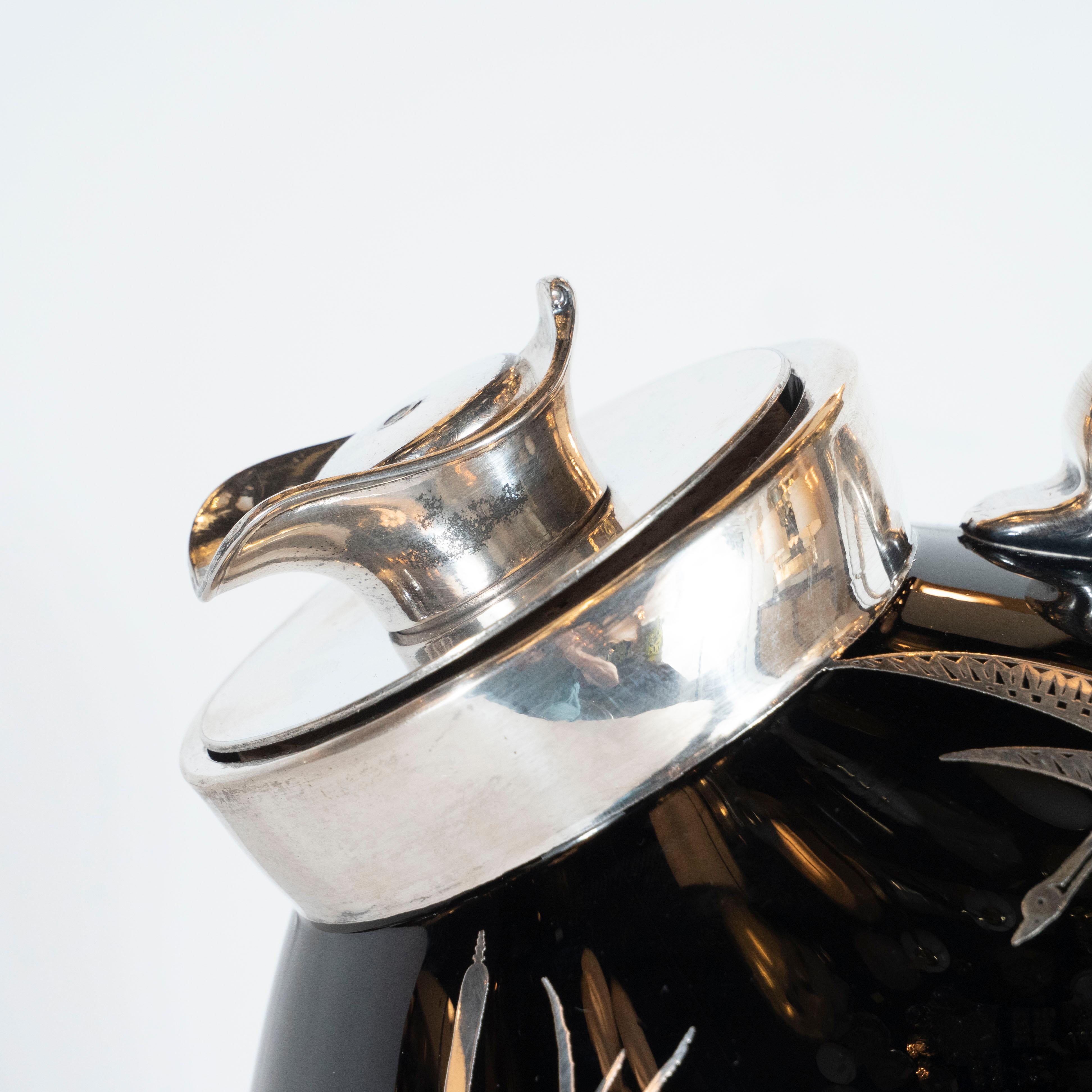 This stunning and dramatic bar pitcher was realized in the United States, circa 1930. A gaggle of stylized geese with geometric motifs composing the interior of their wings flies across the (black glass) sky above an abundance of cattails all