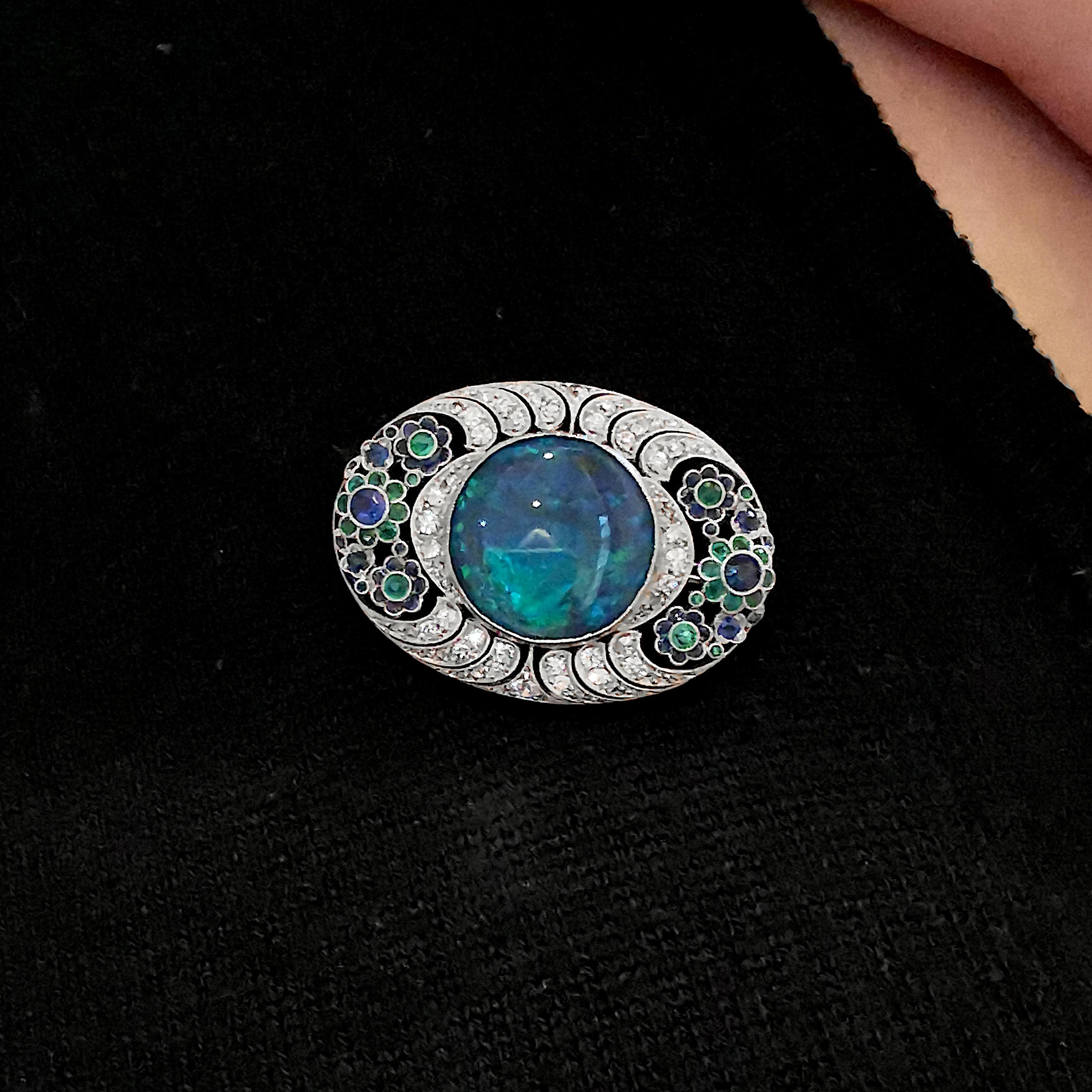An Art Deco opal and diamond brooch, set with a black opal, with blue to green play of colour, in the centre of an oval, rose and eight-cut diamond set, openwork mount, decorated with flowers either side, set with sapphires and emeralds, mounted in