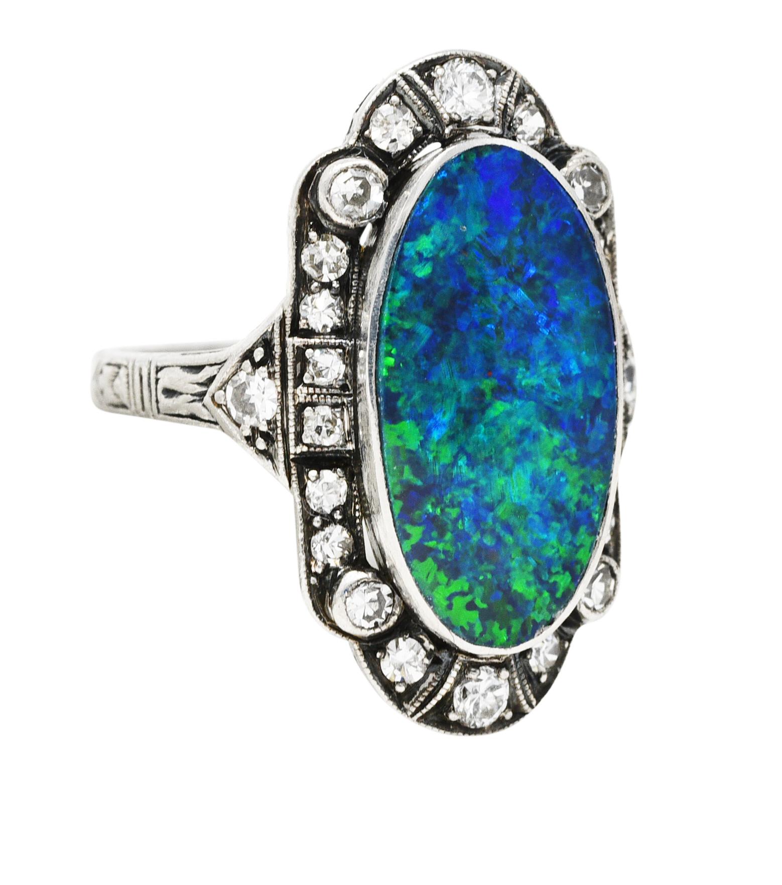 Centering a bezel set oval shaped opal tablet measuring 8.7 x 17.7 mm. Opaque black in body color with strong blue/green play-of-color. With a recessed milgrain surround featuring single cut diamonds. Bead set and weighing approximately 0.32 carat