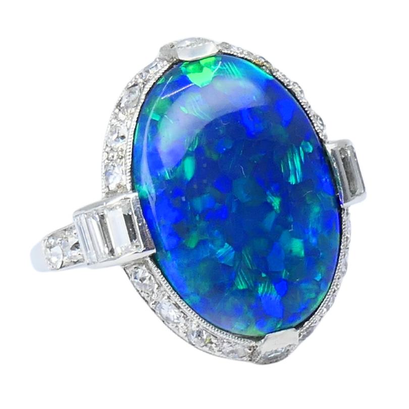 A gorgeous Art Deco black opal ring, made of platinum and featuring diamonds.
The opal displays a harlequin pattern, with vibrant patches of green and blue shifting across the gemstone. The opal is set in a bezel setting with four wide prongs and