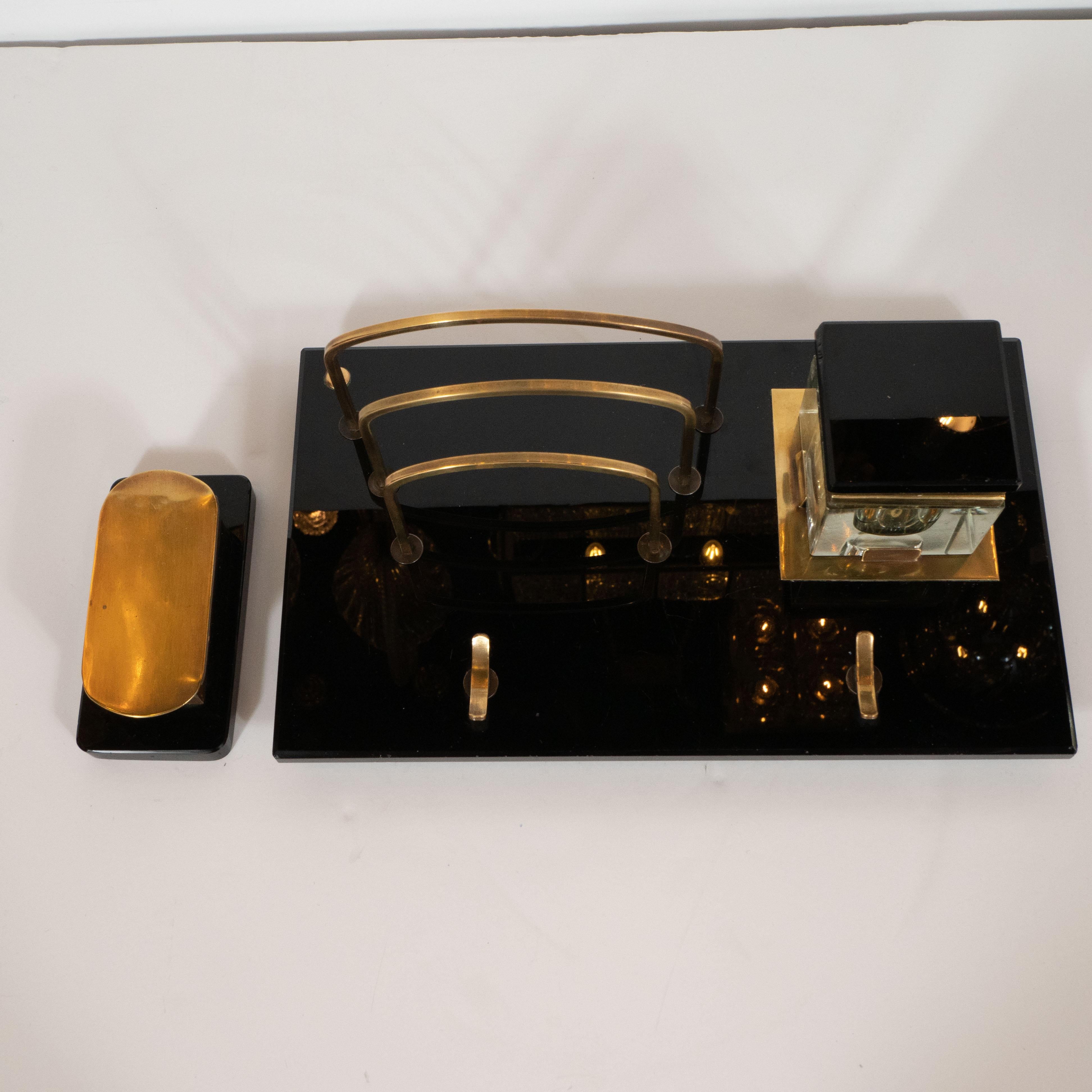 This American Art Deco desk set was produced, circa 1925. The set includes a pen rest, an inkwell, a letter holder and a stamp caddy. The letter holder, inkwell, and pen rest all sit on a lustrous black vitrolite plinth base with beveled edges. The