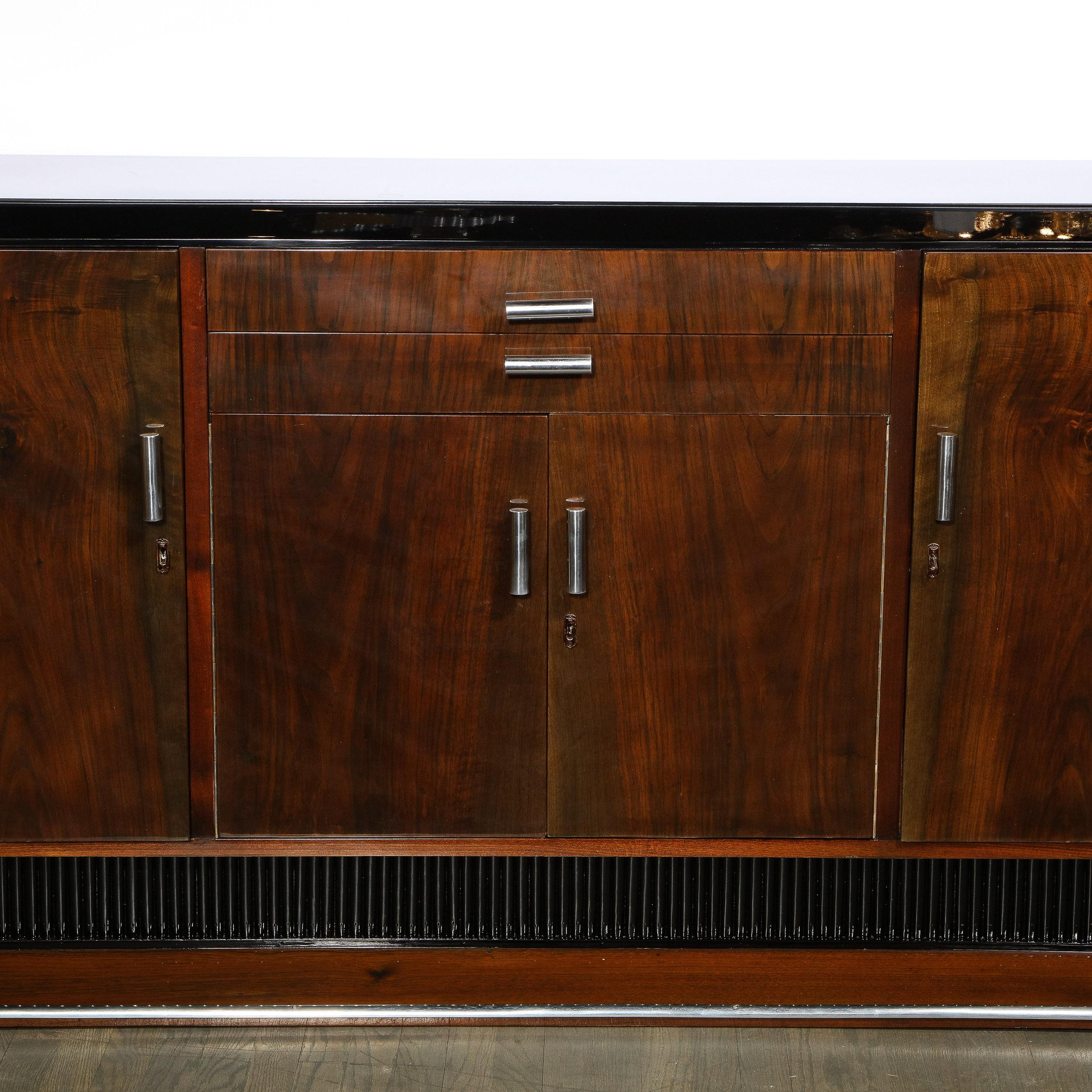 This stunning Art Deco Machine Age skyscraper style sideboard was realized in the United States circa 1935. It features a beveled bookmatched walnut base with an aluminum wrapped perimeter that ascends into a reeded art Deco tier that supports the