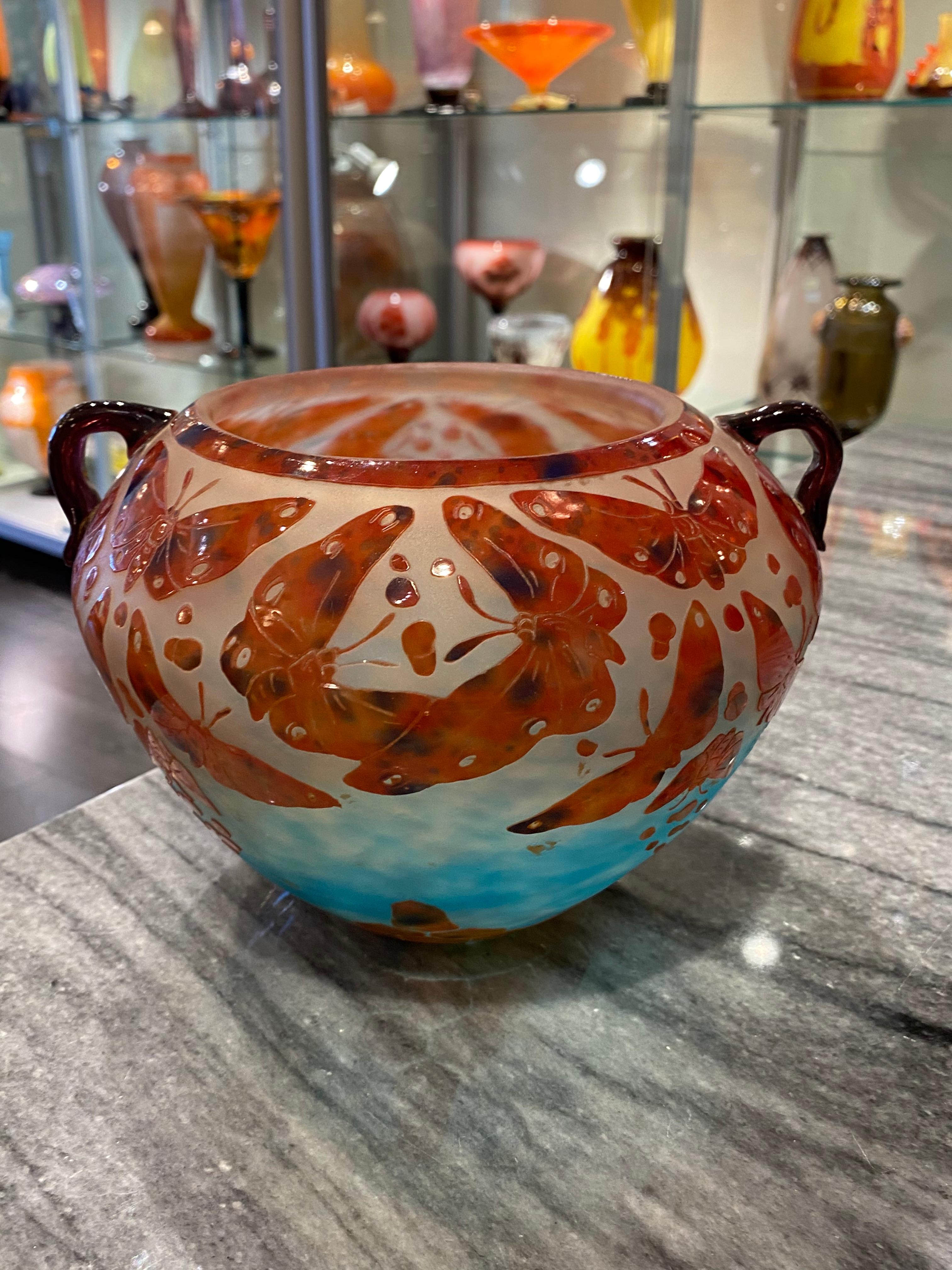 A glass vase in Blue shading into Opalescent White, overlaid with Red speckled with Blue.  The Red Butterflies are acid-etched and spread through out the vase.  There are handles (Violet color) on this vase on each side.  This piece is part of the