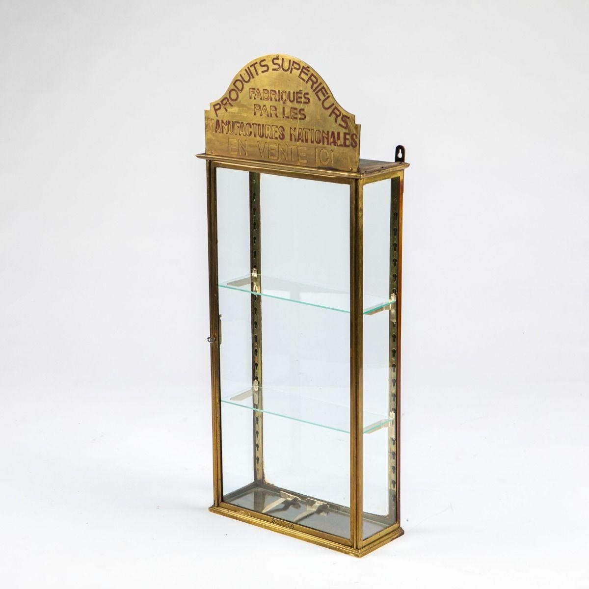 Vitrine décorative Art Déco en laiton avec verre sur les quatre côtés. Les étagères en verre réglables sont montées avec des supports en laiton nouvellement fabriqués dans le style des originaux. Au sommet, un panneau en laiton avec un texte gravé