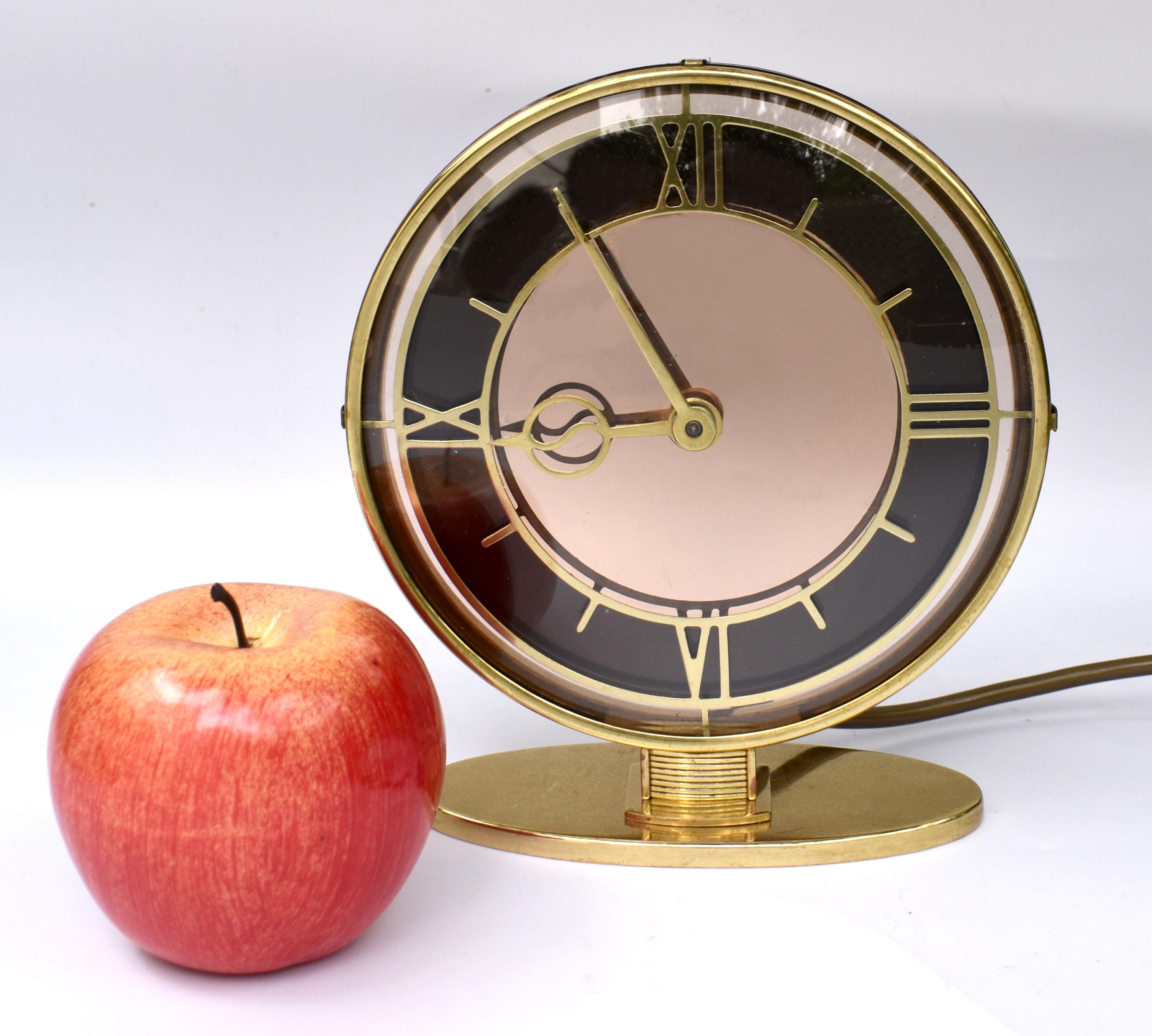 Nous vous proposons cette horloge électrique Art Déco, de fabrication britannique et datant des années 1930. Le cadran est composé d'un verre transparent et d'un miroir rose pâle avec une lunette en laiton et des chiffres stylisés. L'ensemble de