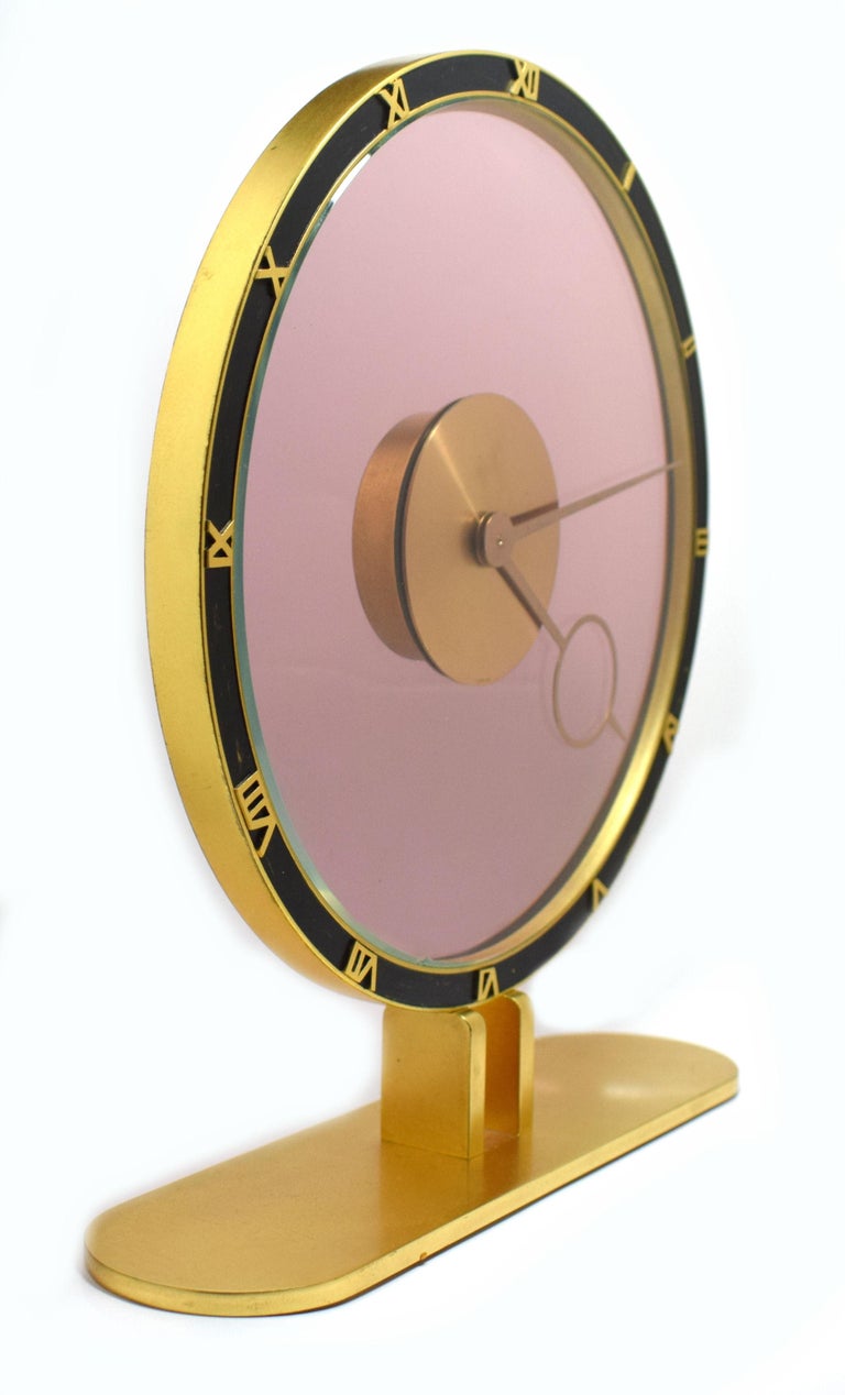 Superbly stylish is this wonderful Art Deco 8 day clock by Kienzel. Beautiful rose colored glass and brass desk timepiece is typical of the 1930s.
The brass case is polished and lacquered. The chapter ring with raised brass Roman numerals on a