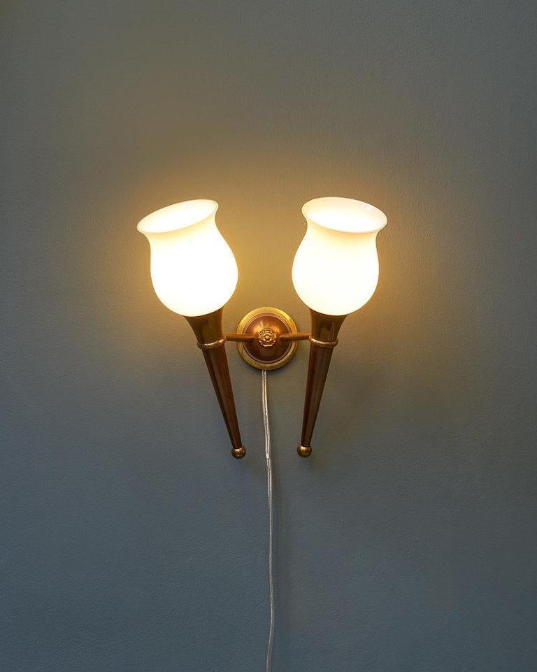 A stunning pair of bronzed wall sconces from France with double torch-shaped arms, opaque glass tulip shades, and a floral medallion. Similar in style to Maison Jansen lighting. France, circa 1940s. These are hardwired and ready for use.