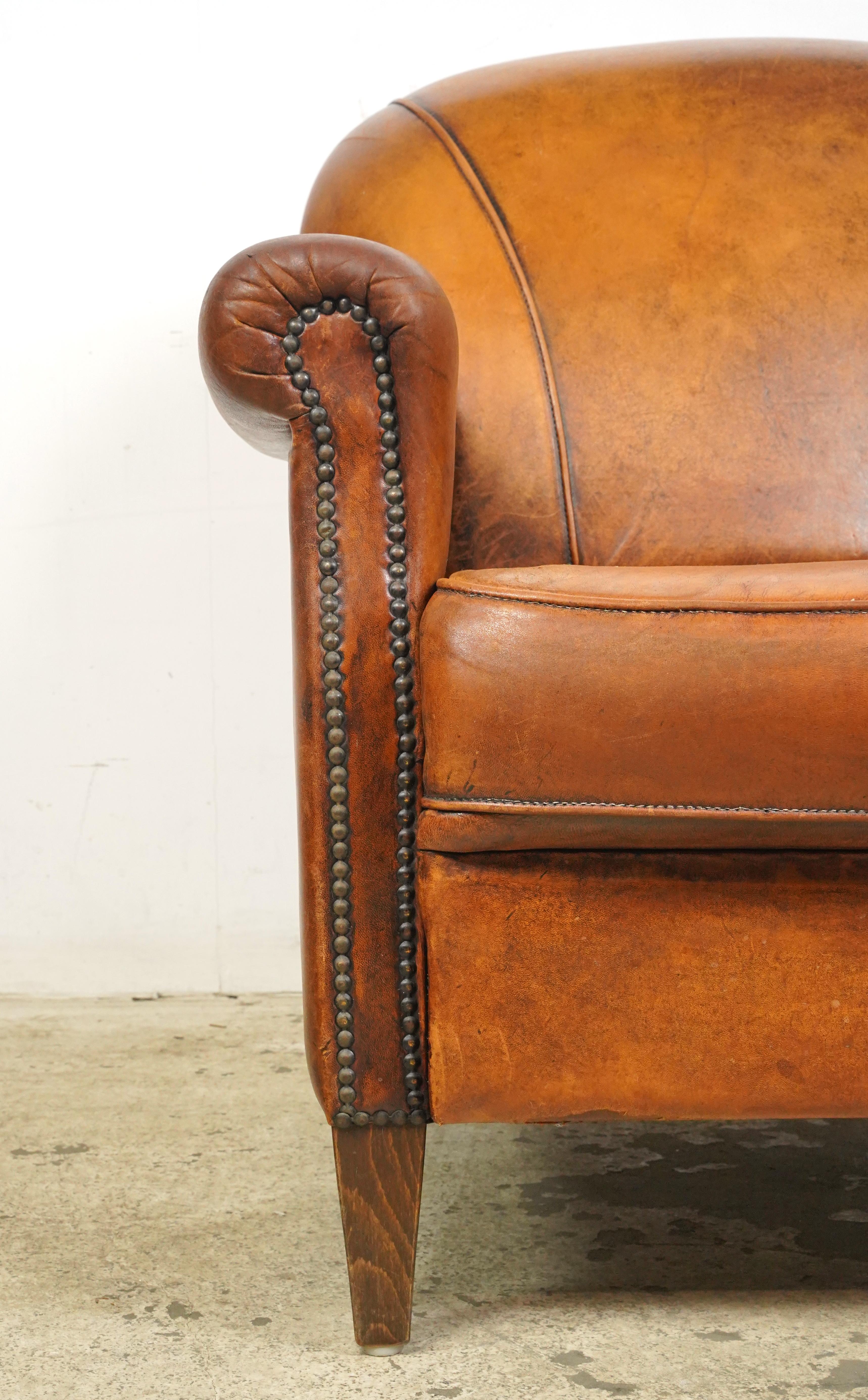 Art Deco style dark brown leather studded club chair made in France. It has a rounded back, rolled armrests, a cushioned seat, studded details, and wooden tapered legs. The leather and feet show signs of wear and age. Please refer to the pictures