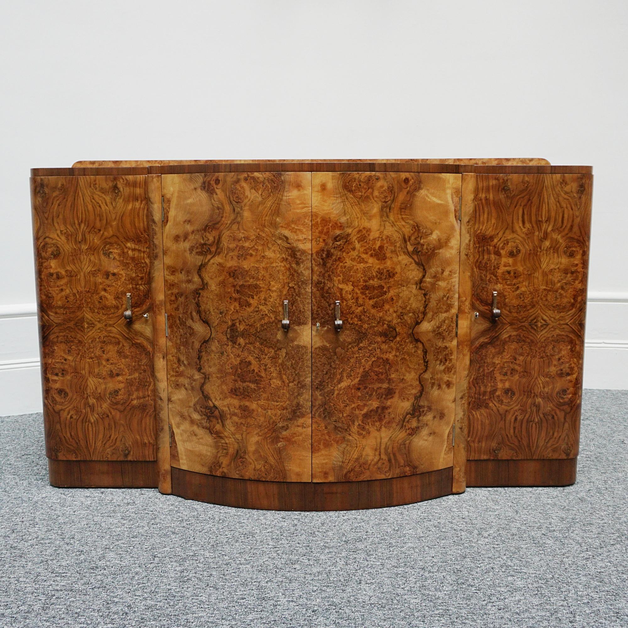 An Art Deco sideboard by Harry & Lou Epstein. Burr walnut veneer with figured walnut banding To either side interior shelved compartments, with central section opening to reveal three drawers and lower storage compartment. Original walnut and brass