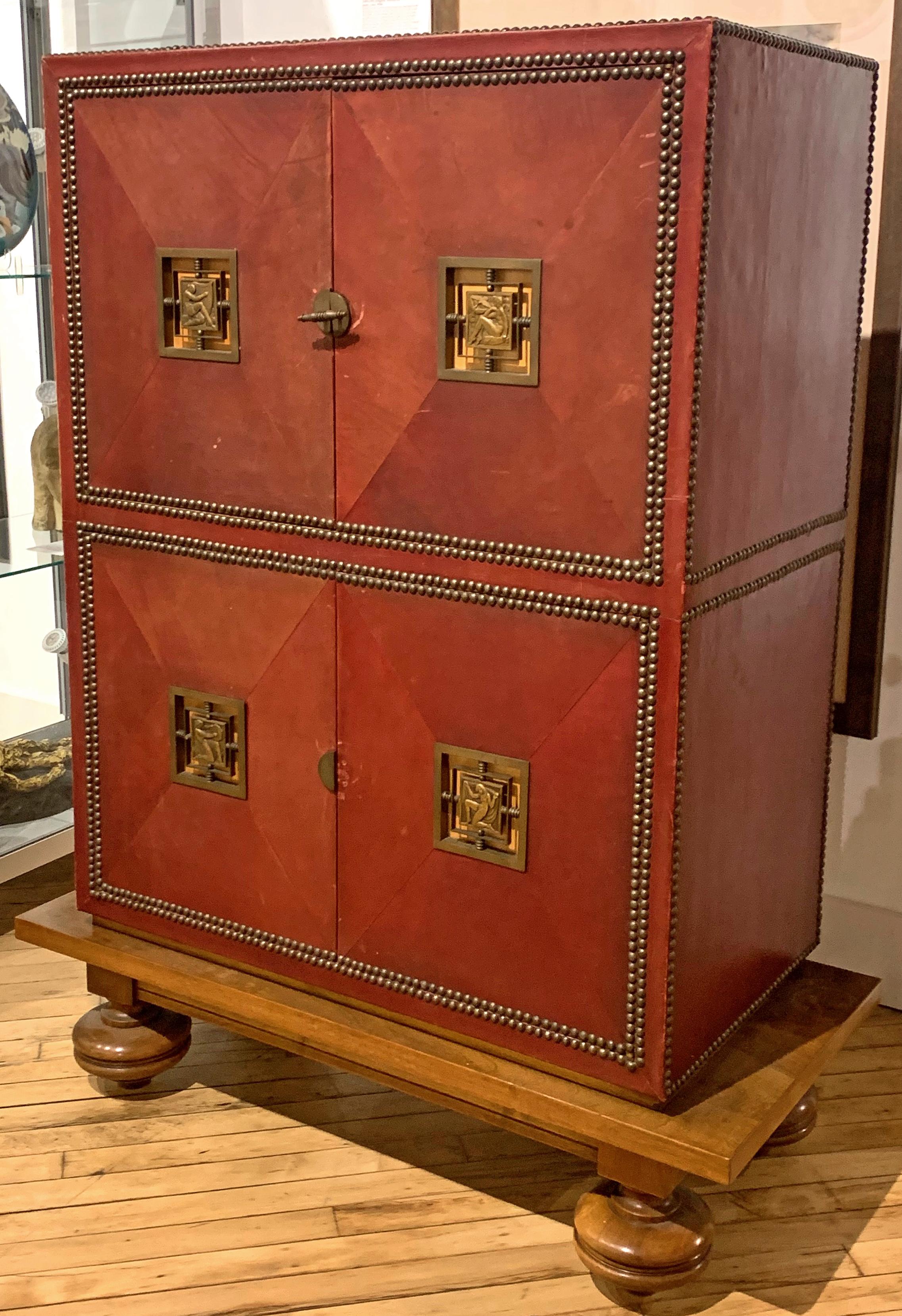 Beautifully proportioned, constructed and finished, this unique and remarkable Art Deco cabinet is covered with leather dyed a rich, deep red color, and features four Art Deco bronze mounts depicting nude figures celebrating Music and Art. The